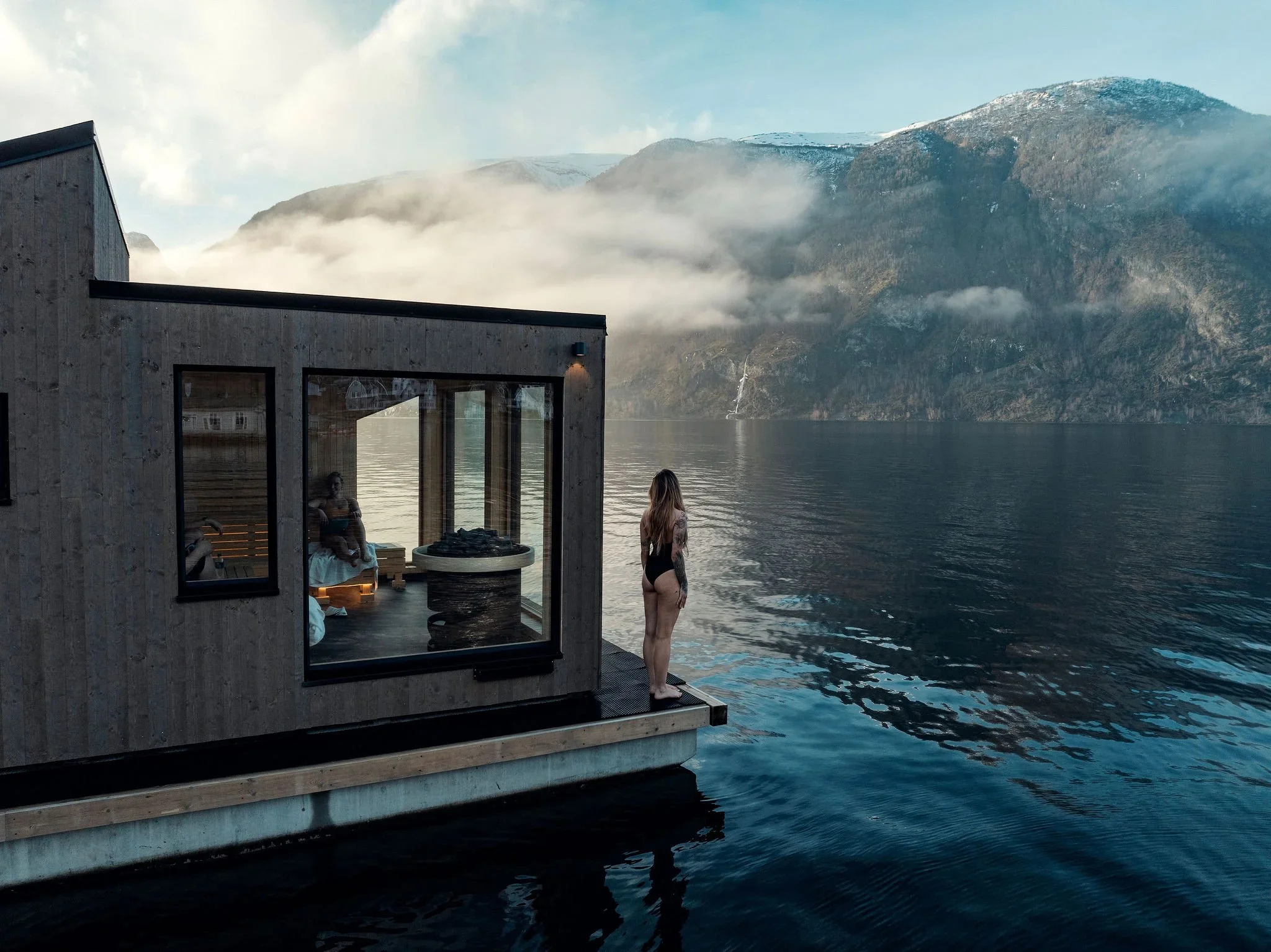 A woman stands outside a modern floating sauna overlooking calm fjord waters and mist-covered mountains in Norway.