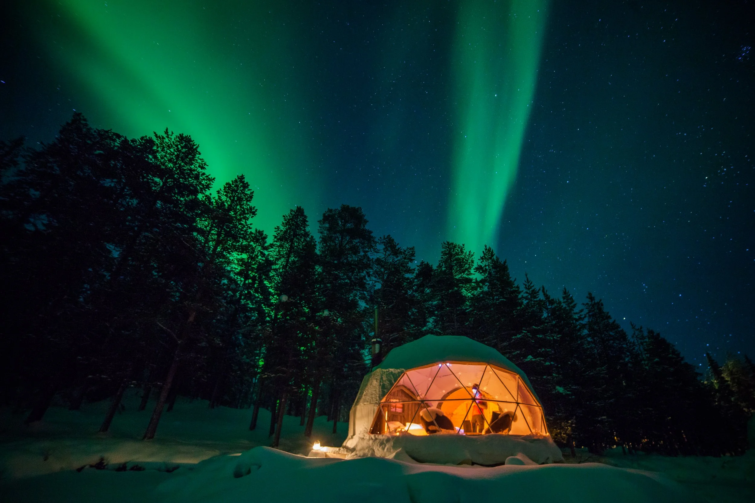 Glass-fronted aurora dome glowing in a snowy forest beneath the Northern Lights in Torassieppi, Finnish Lapland.