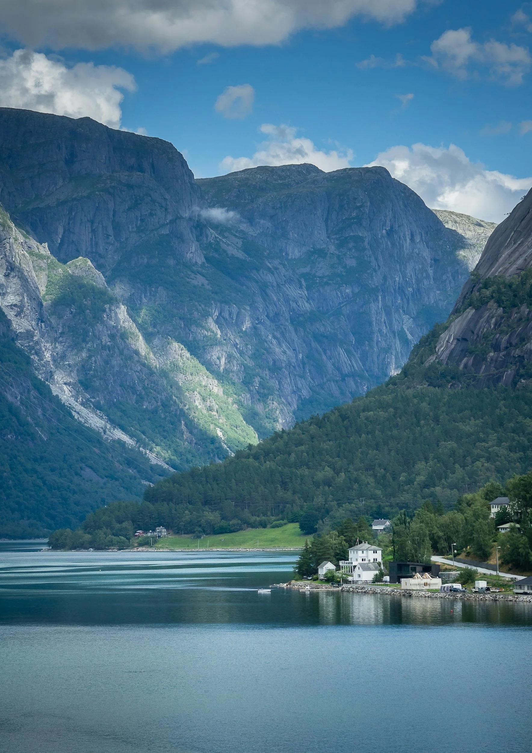 Small coastal village beside a tranquil Norwegian fjord, surrounded by steep, forested mountains beneath a blue summer sky.
