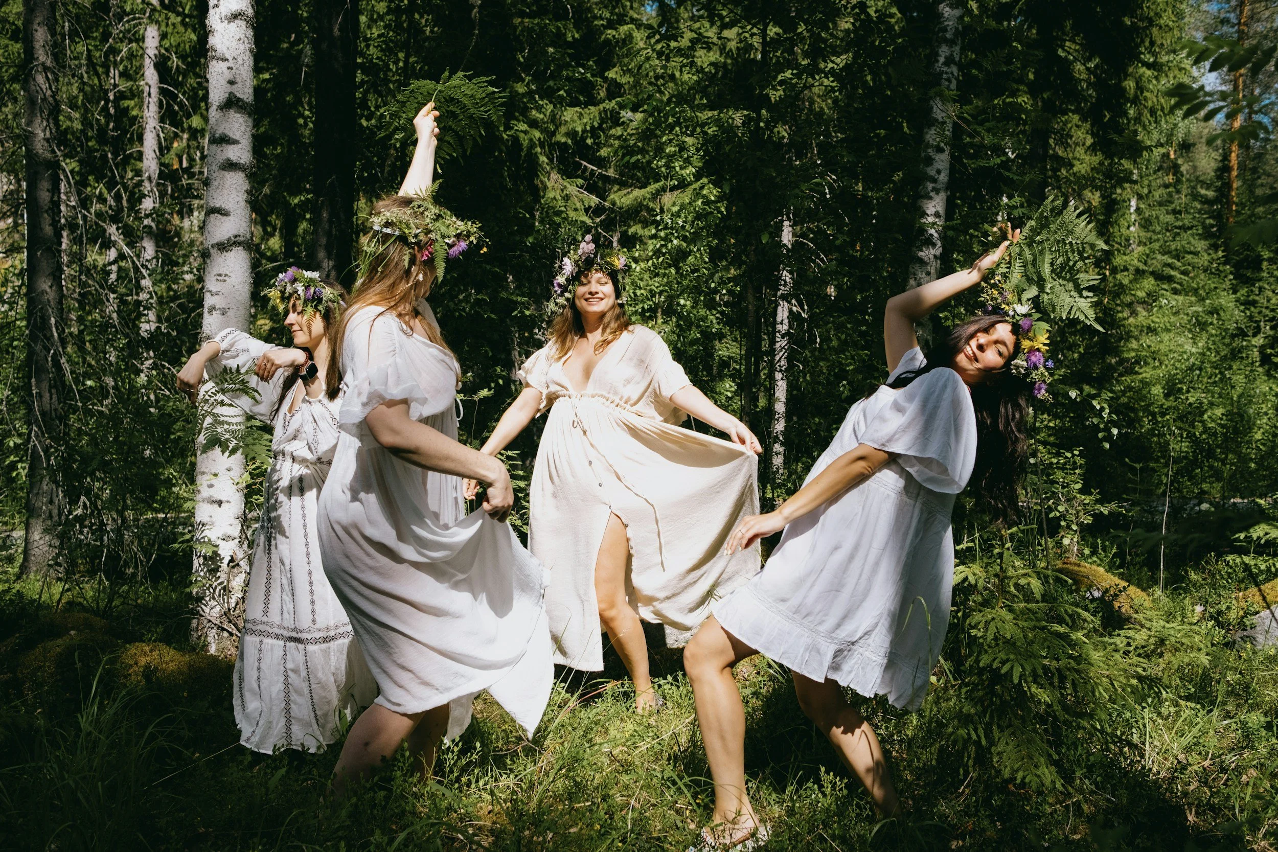 Four women wearing white dresses and flower crowns celebrate Finnish Midsummer in a sunlit forest.