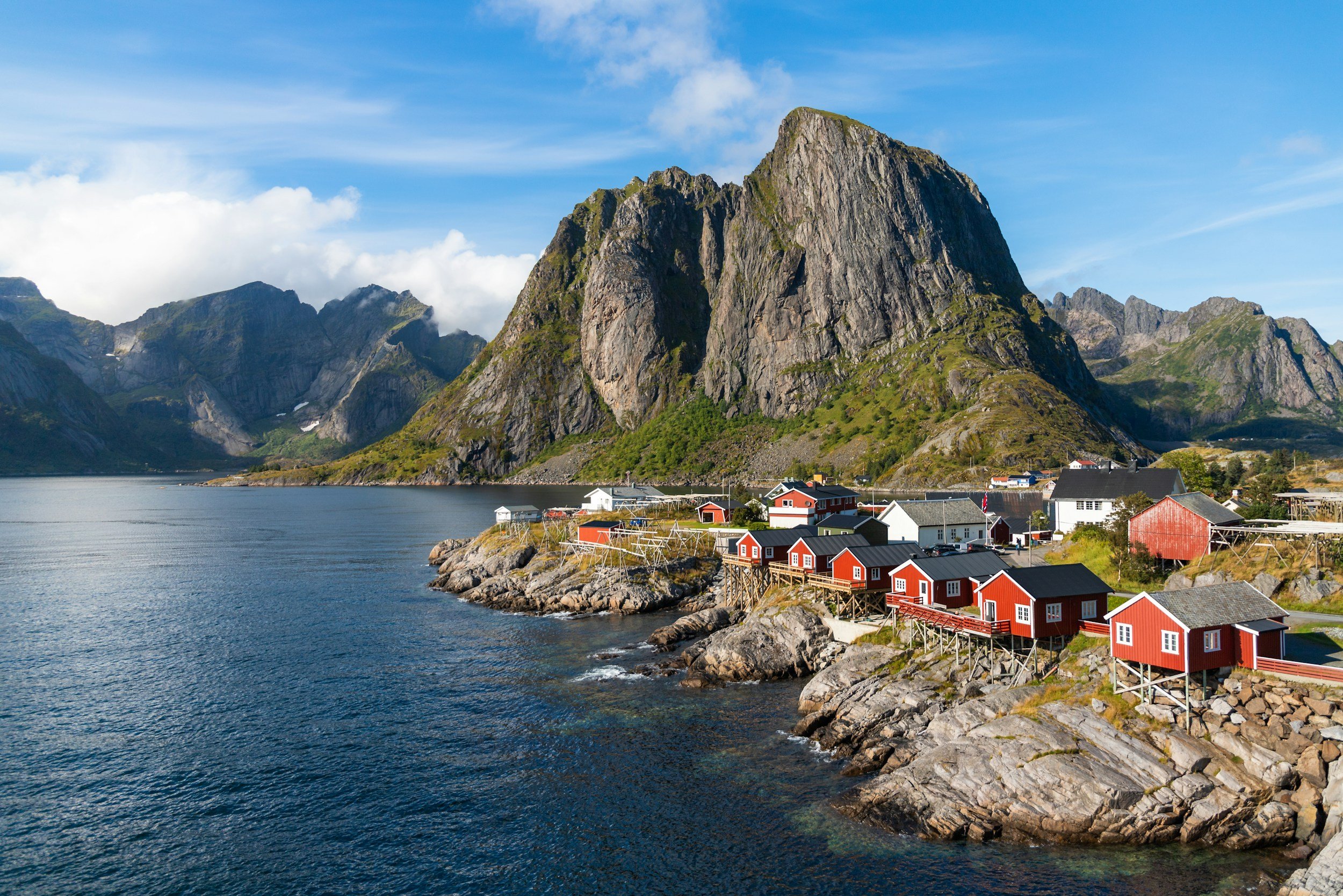 Red fishing cabins beside dramatic mountains and clear coastal waters in the Lofoten Islands, Norway.