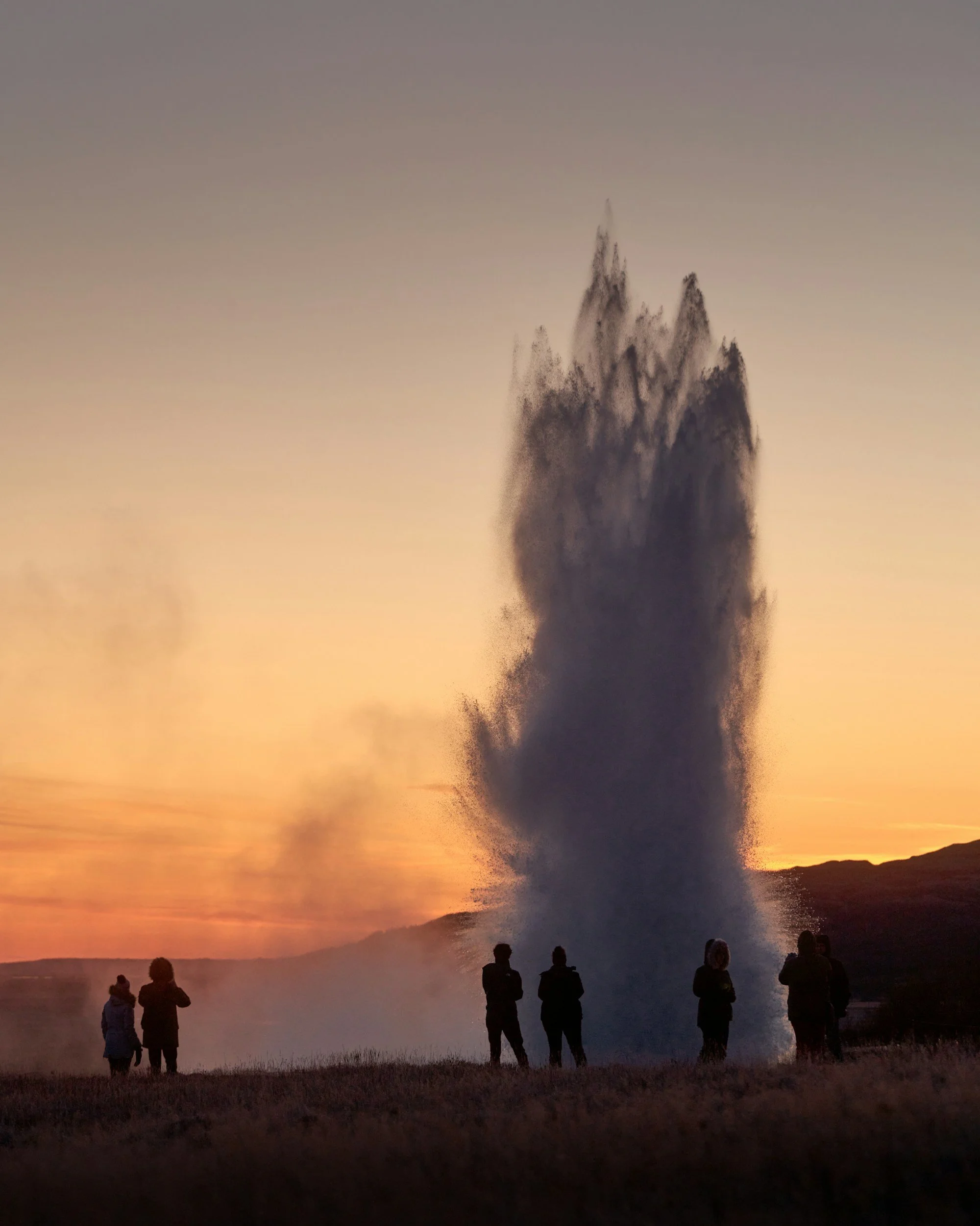 Visitors watching a powerful geyser erupt against a dramatic sunset sky in Iceland’s Golden Circle.