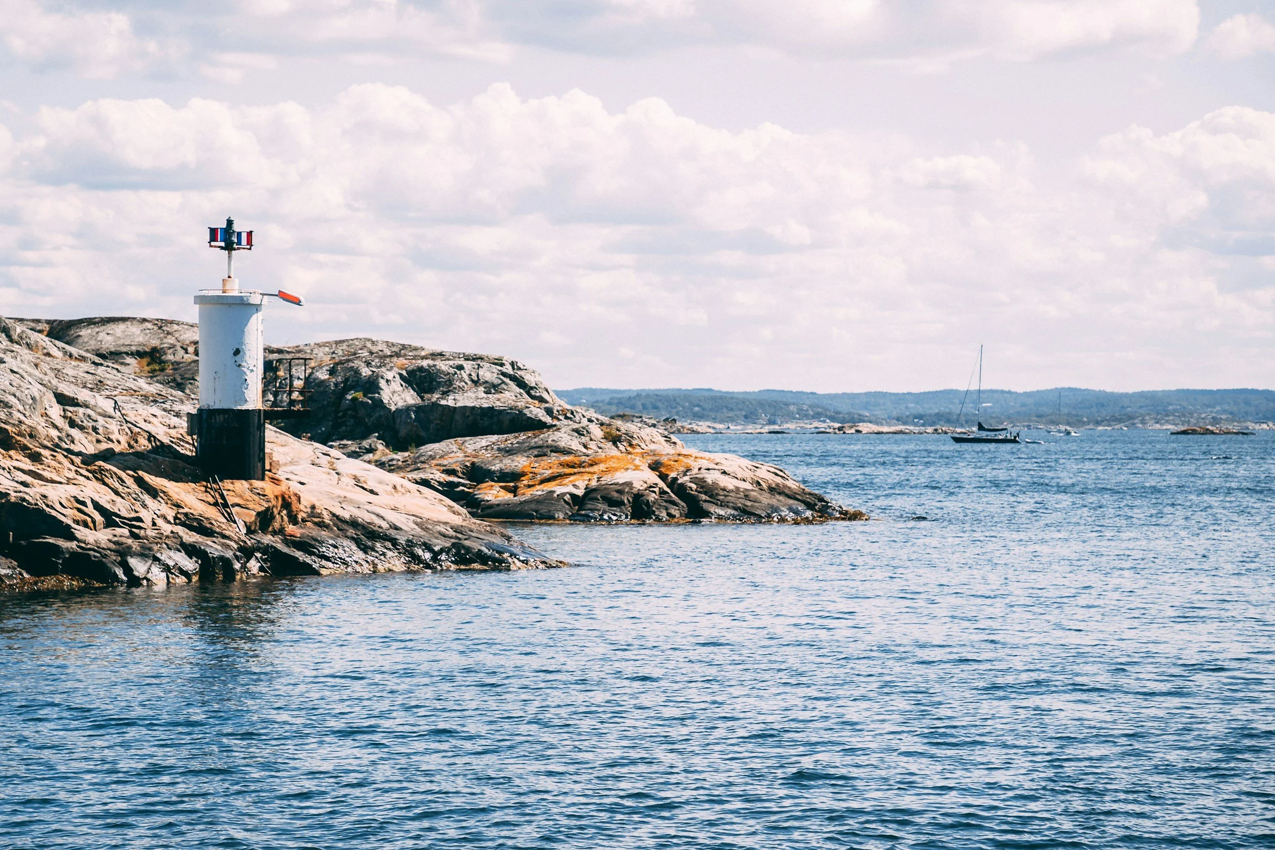 Small lighthouse on a rocky island with sailing boats crossing the calm waters of the Swedish archipelago.
