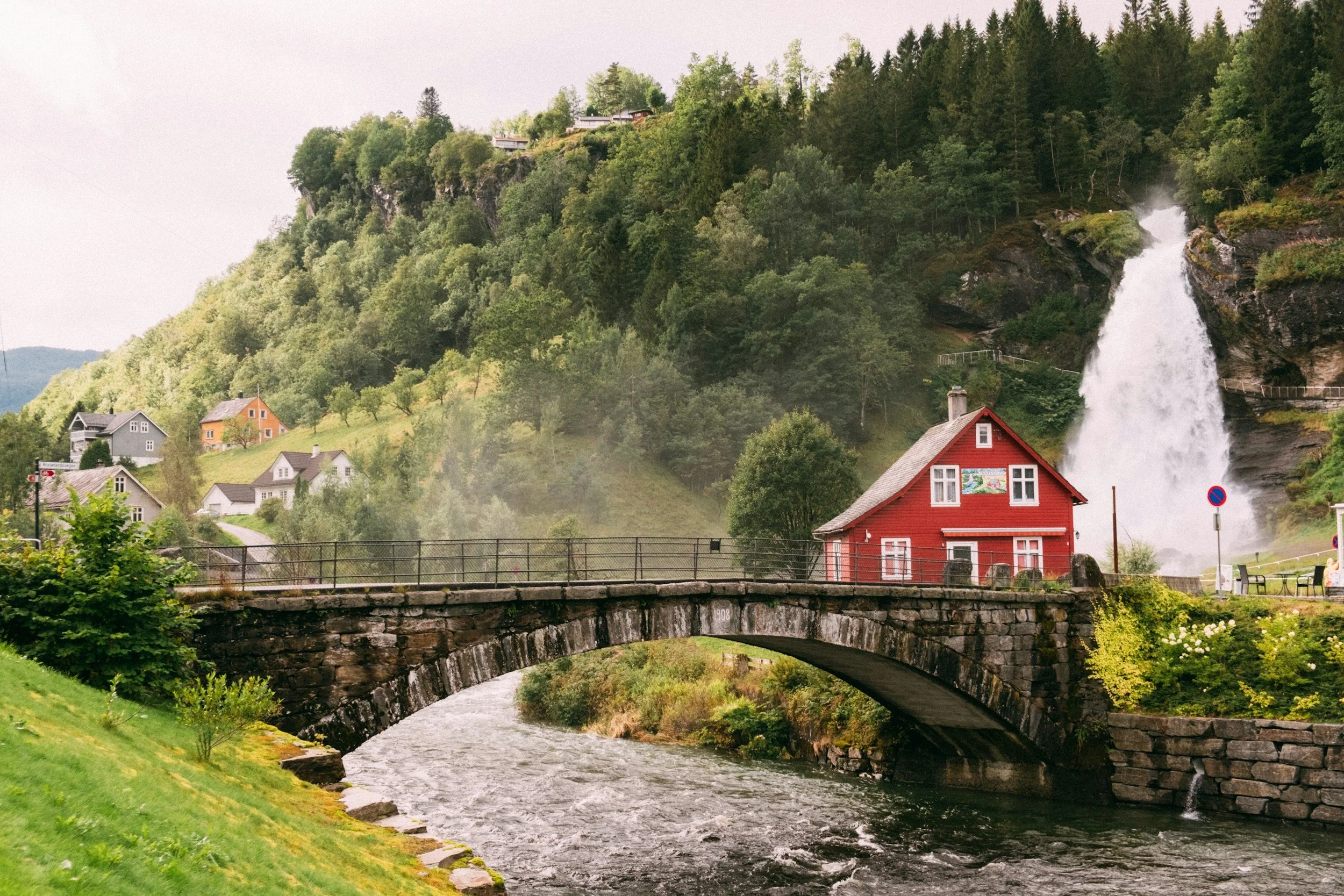 Powerful waterfall beside a red wooden house and historic stone bridge in rural Norway.
