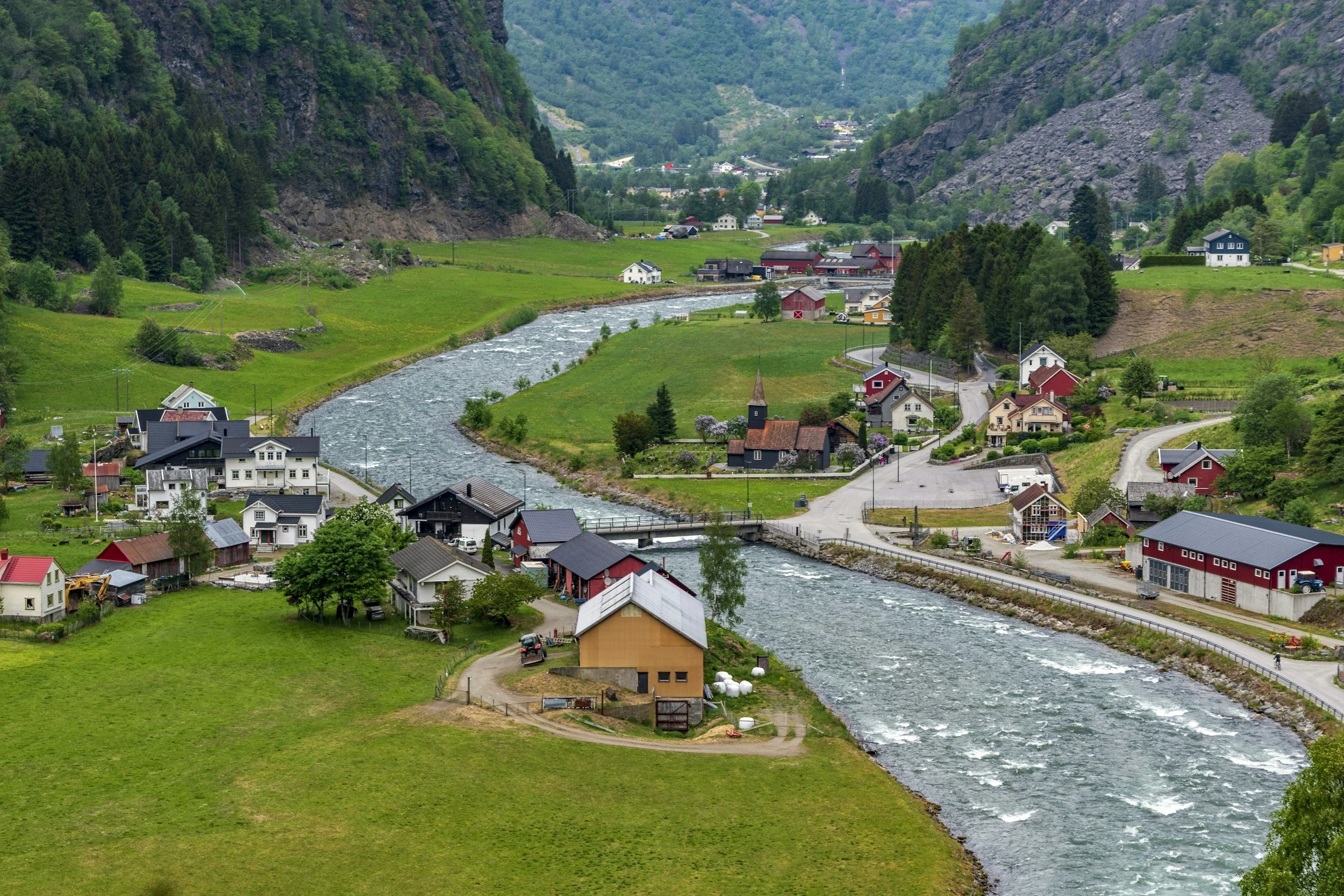 Traditional village and winding river surrounded by green fields and steep mountains in rural Norway.