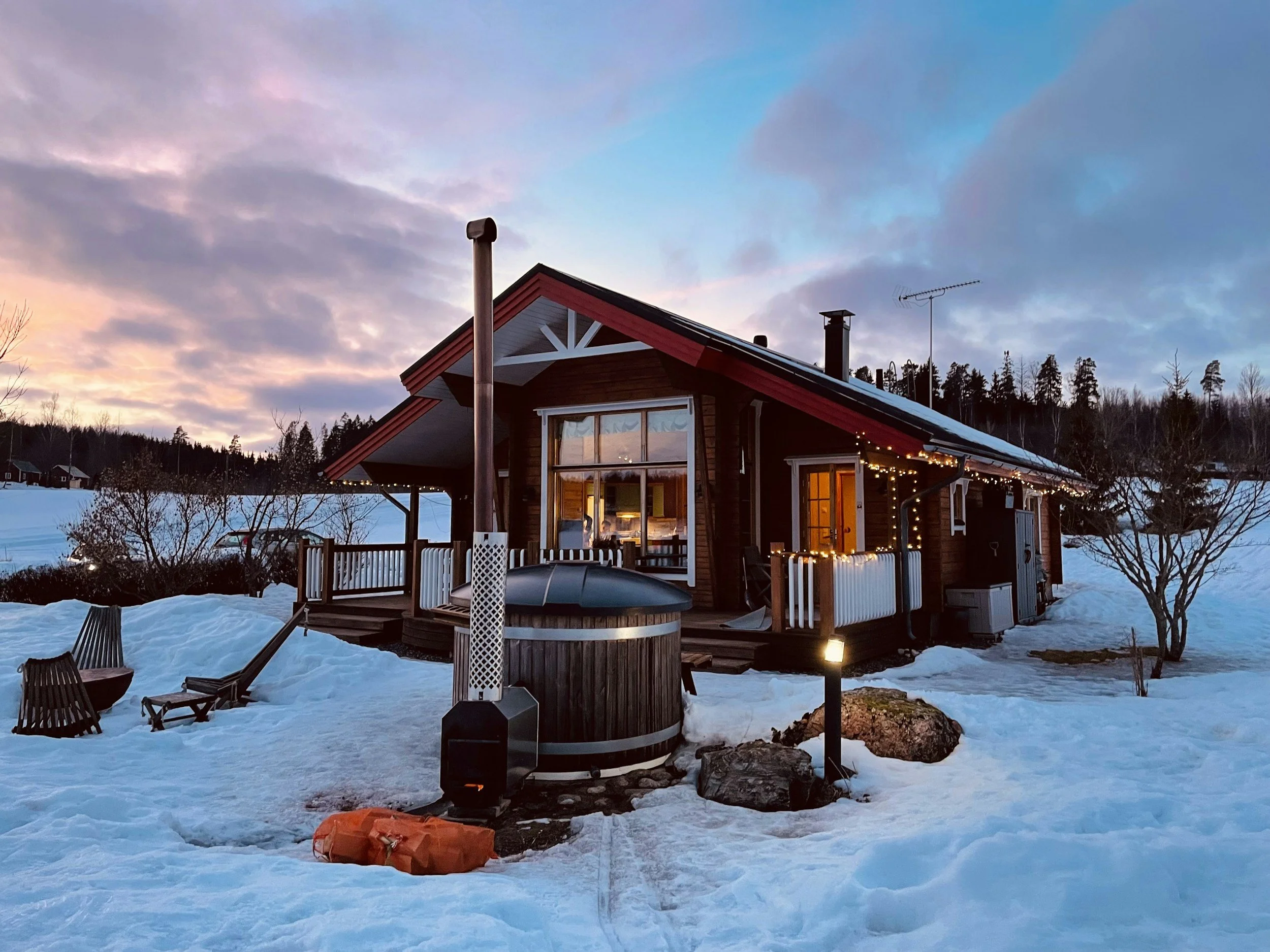 Snow-covered wooden cabin with an outdoor hot tub at sunset in Karjalohja, Finland.