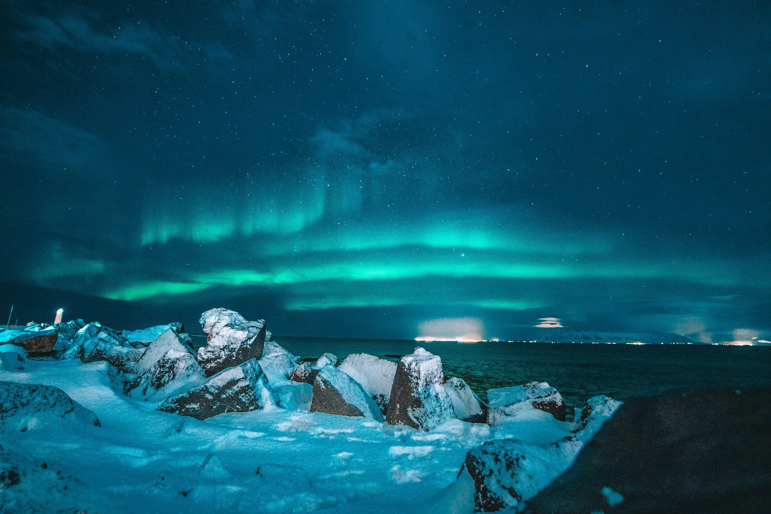 Green Northern Lights stretch across a star-filled sky above a snowy rocky coastline in Iceland.