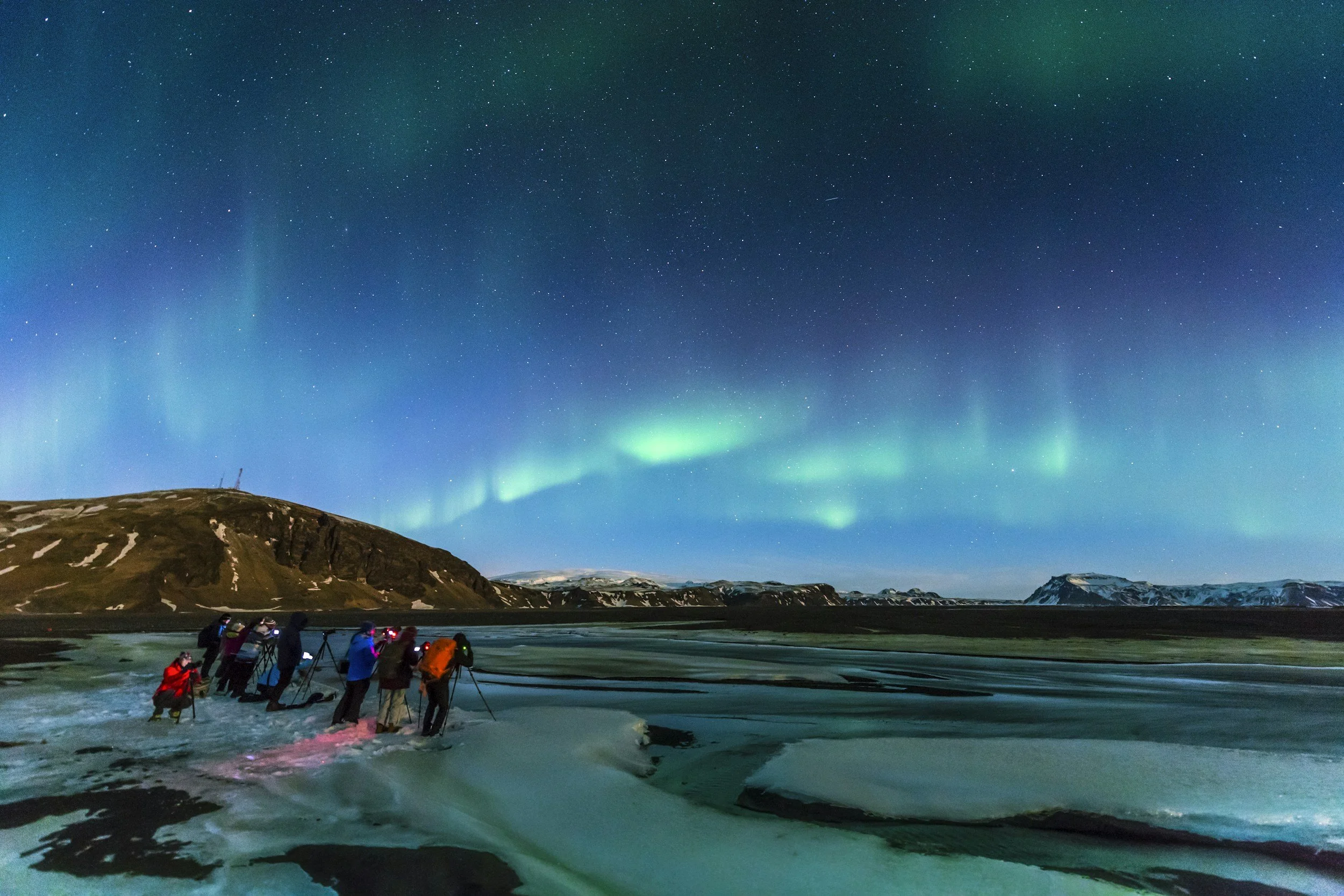 Photographers set up tripods beneath green Northern Lights over a wintry Icelandic landscape.