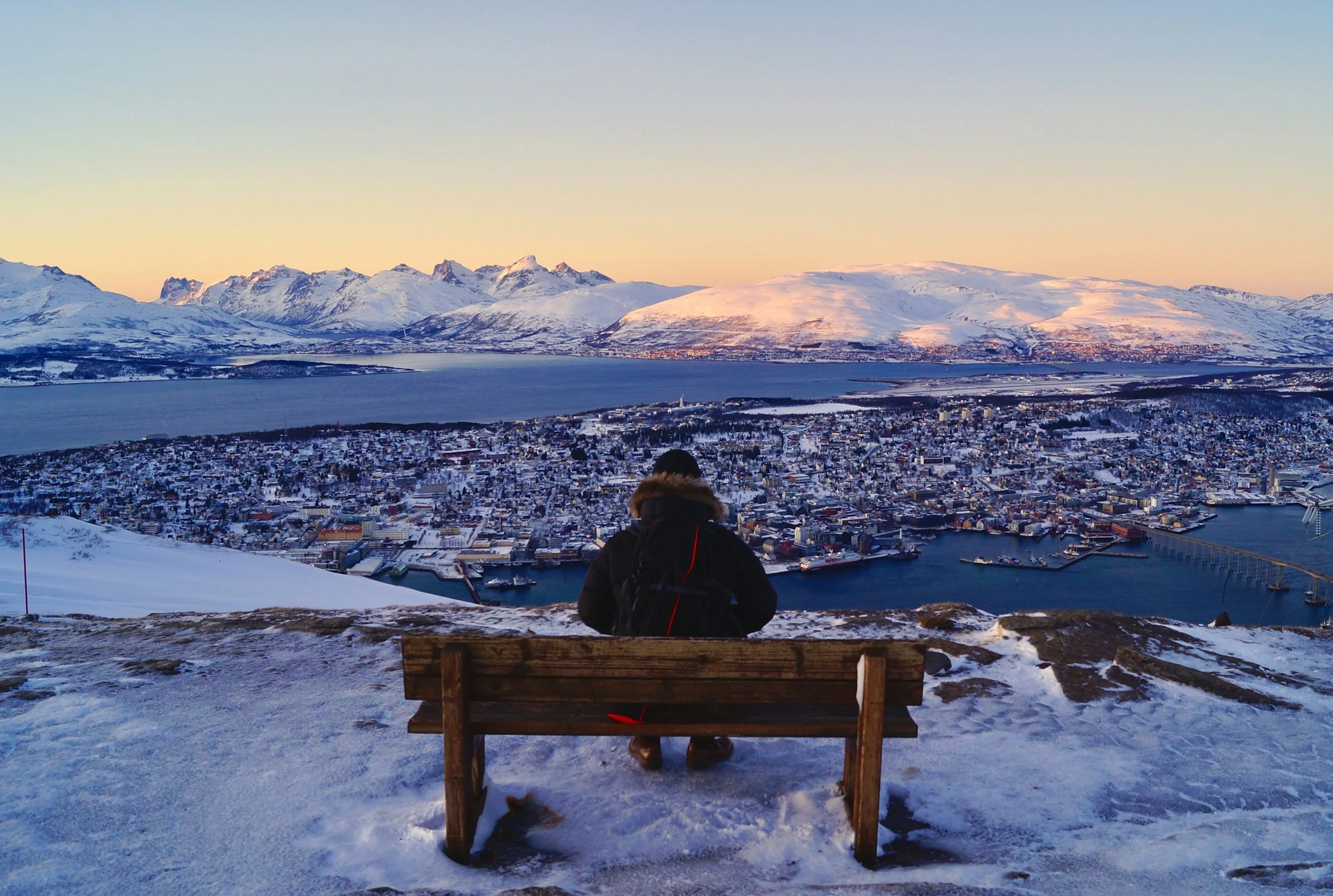 A traveller sits on a bench overlooking snowy Tromsø, its fjords and surrounding mountains in winter.