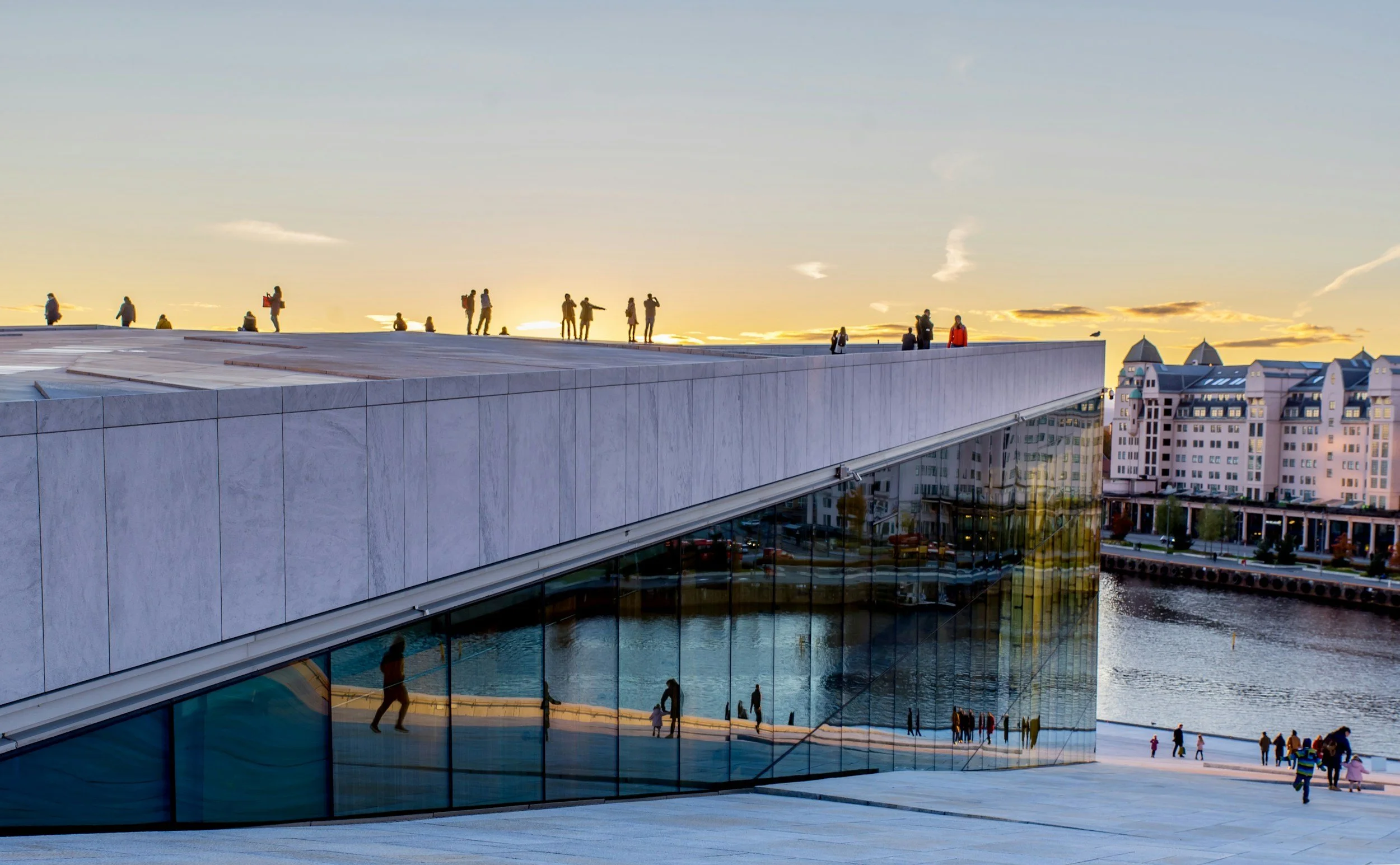 Visitors walk across the sloping roof of Oslo Opera House beside the waterfront at sunset.