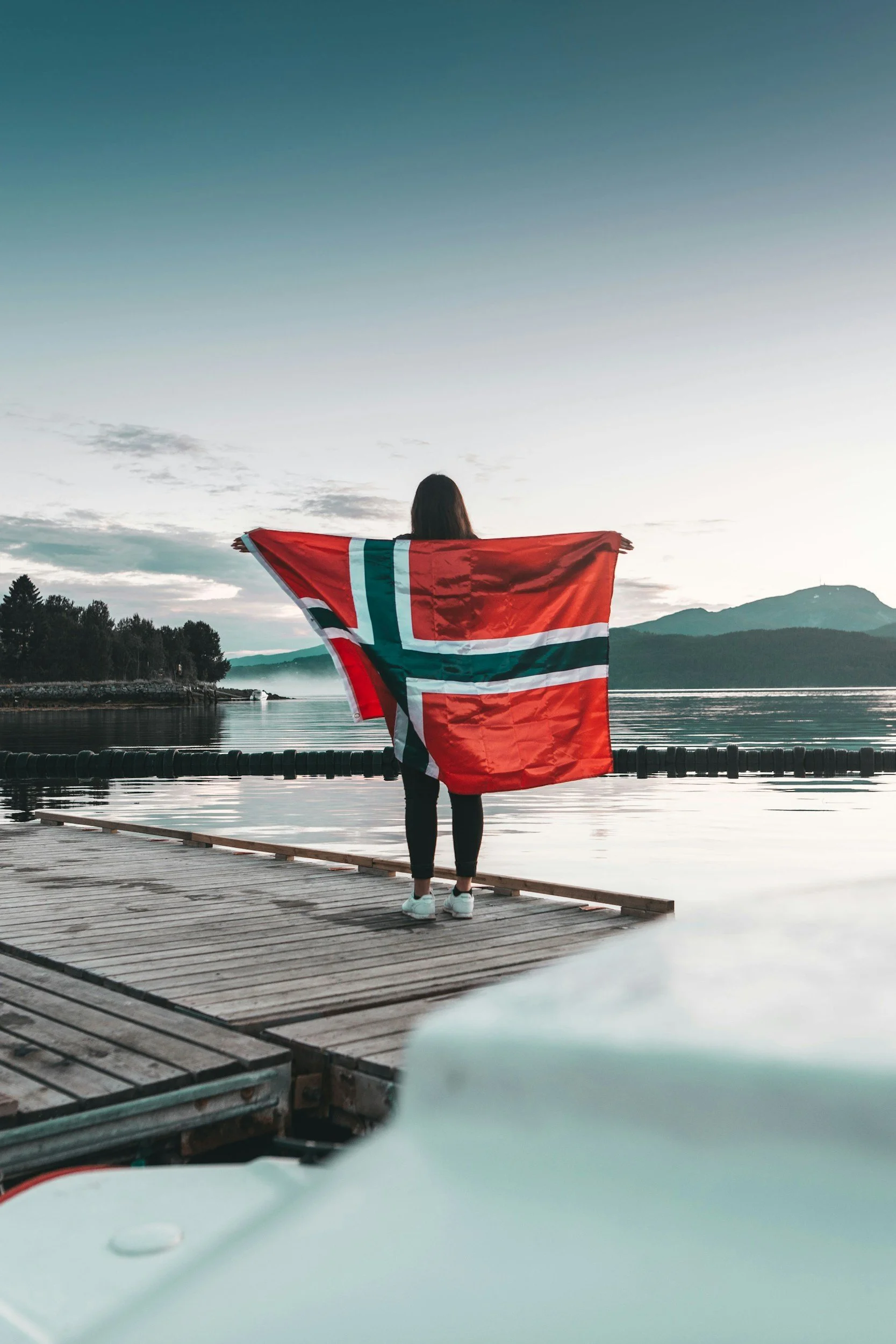 A woman holds a large Norwegian flag while standing on a waterfront dock overlooking mountains and calm water in Norway.
