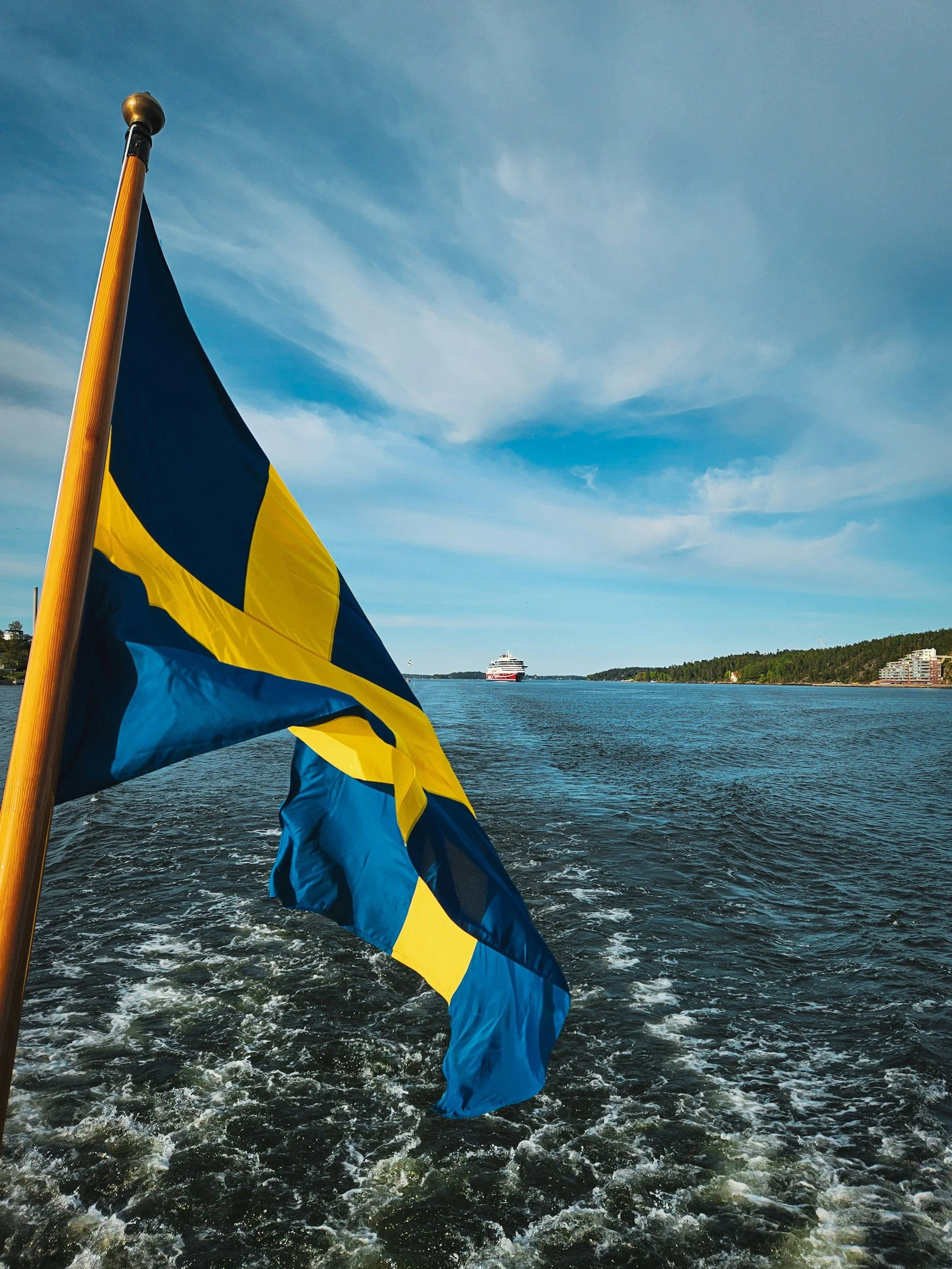 A Swedish flag flies from a boat travelling across blue water with islands in the distance.