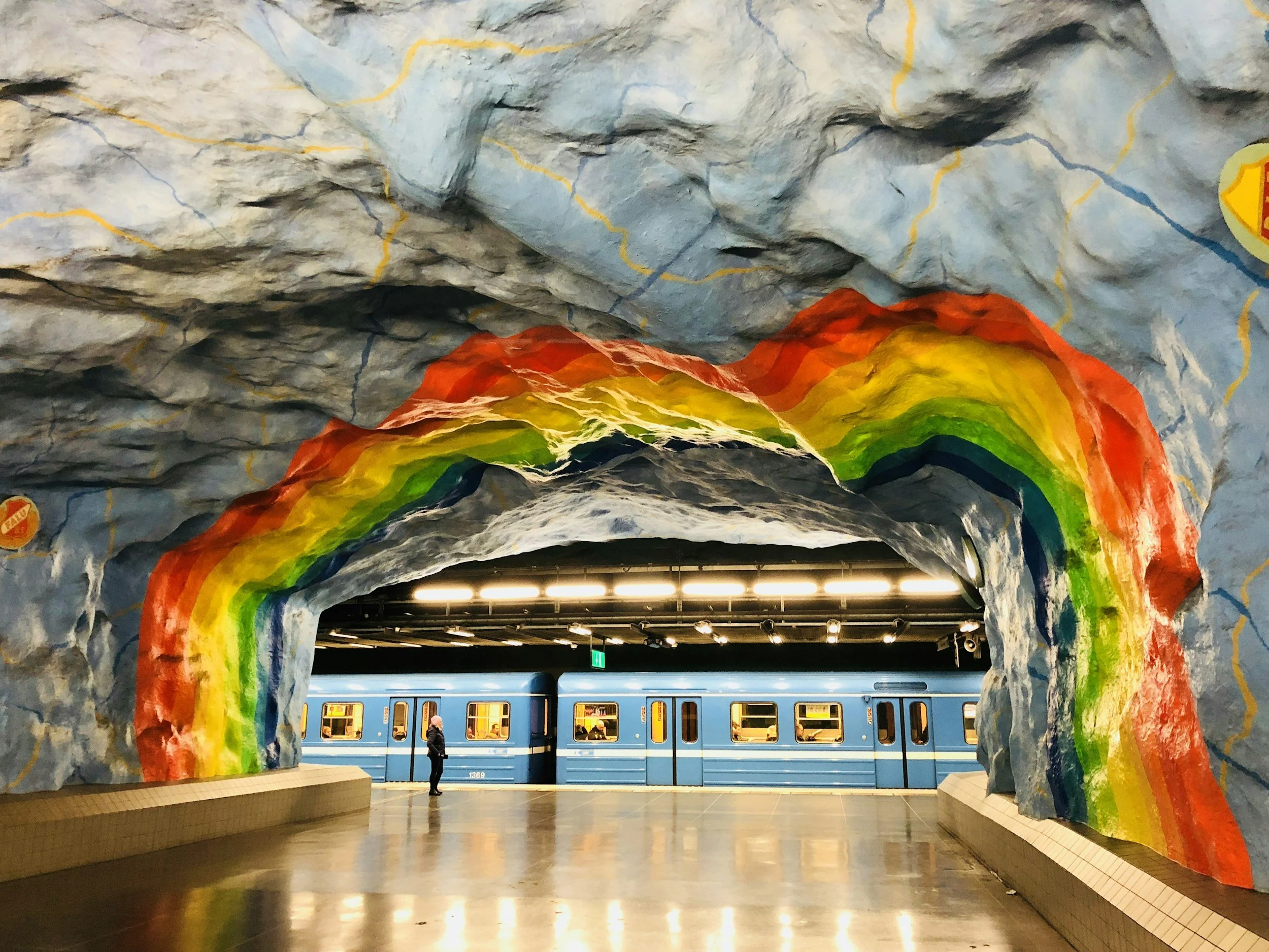 A blue Stockholm Metro train stands beside a colourful rainbow painted across a cave-like underground station.