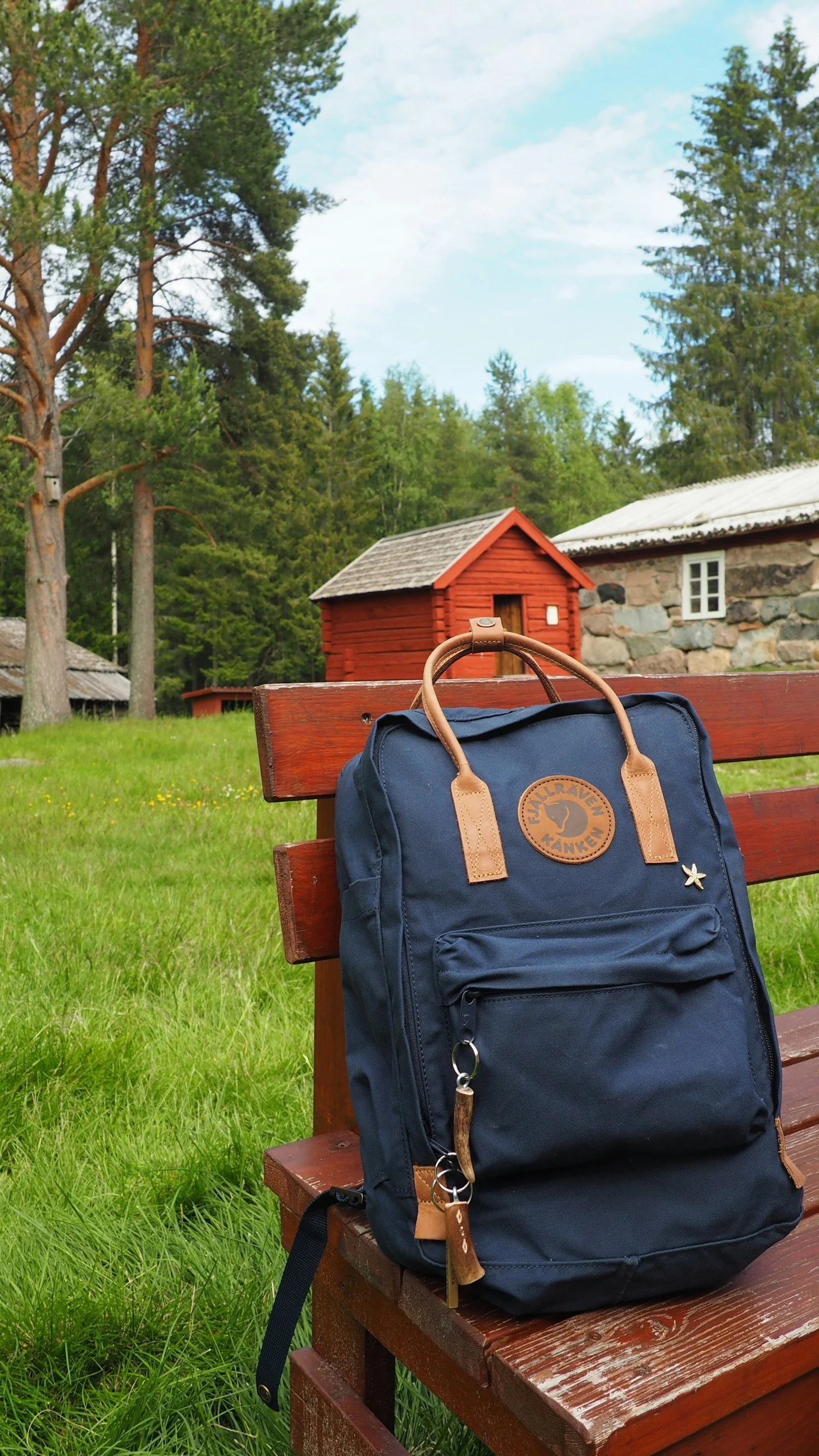 A blue Fjällräven backpack rests on a wooden bench beside traditional red cottages and pine trees during Midsummer in Sweden.