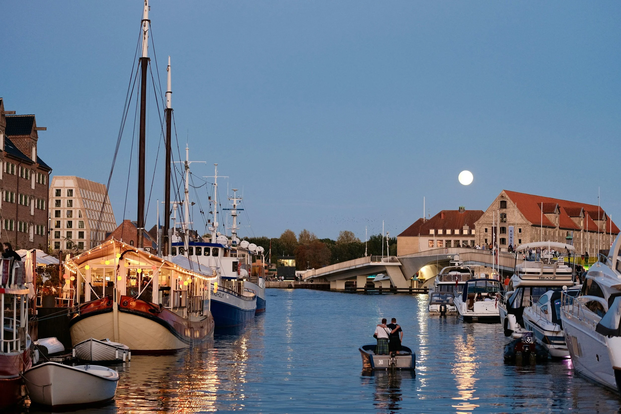 Boats sit along Copenhagen’s Nyhavn harbour beneath a full moon in the evening sky.