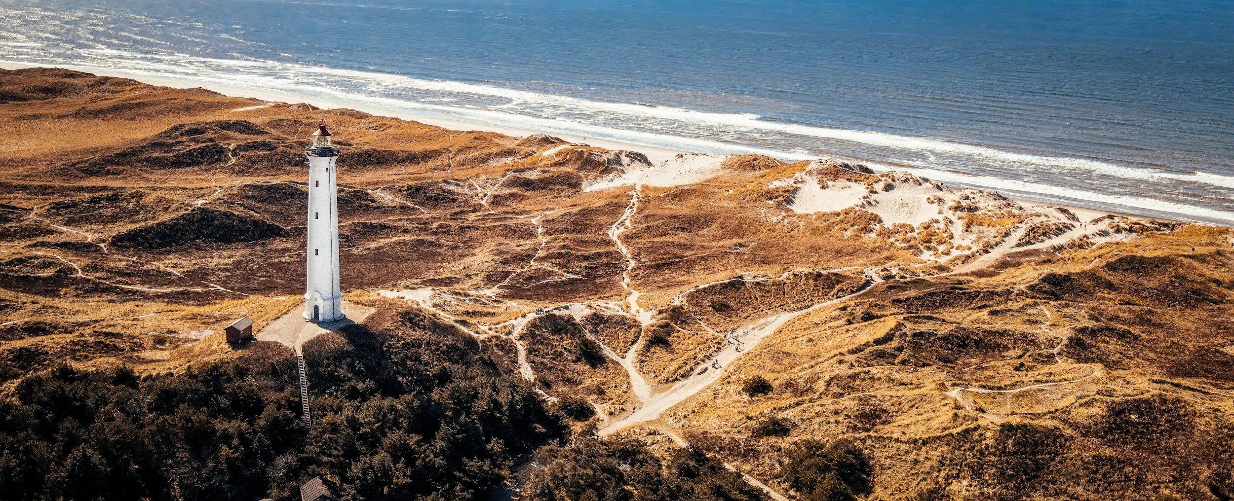 A tall white lighthouse rises above sandy coastal dunes beside the North Sea at Hvide Sande, Denmark.