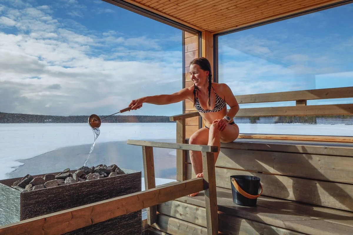 A woman pours water onto hot sauna stones inside a lakeside sauna overlooking a frozen Finnish landscape.