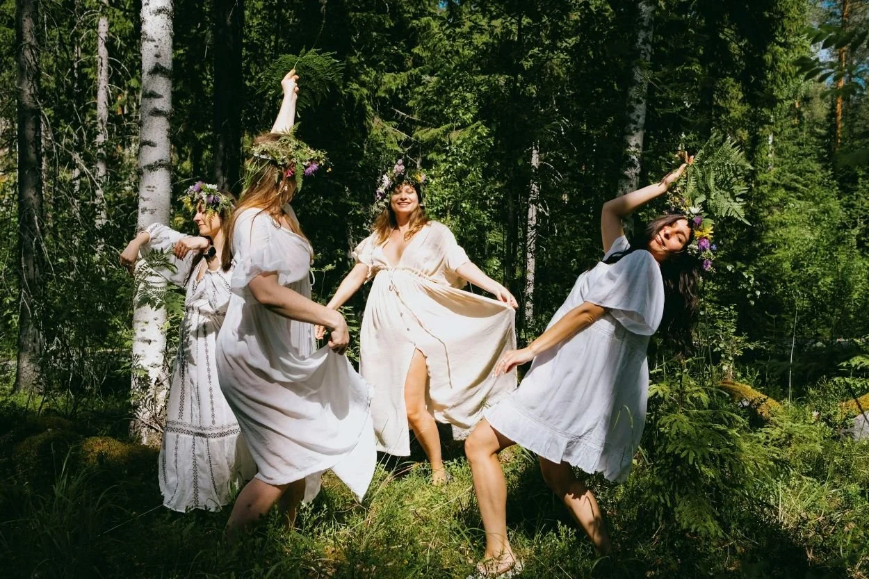 Four women wearing white dresses and flower crowns celebrate Finnish Midsummer in a sunlit forest.