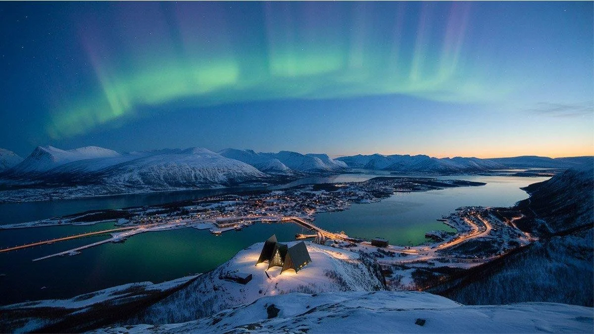 Northern Lights glowing above snow-covered Tromsø, its surrounding fjords and mountains, viewed from Fjellheisen in winter.