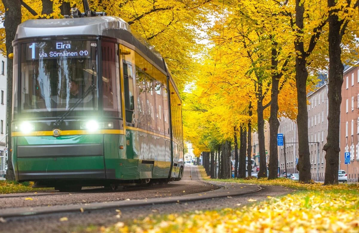 A green Helsinki tram travels through Sörnäinen beneath trees filled with bright yellow autumn leaves.