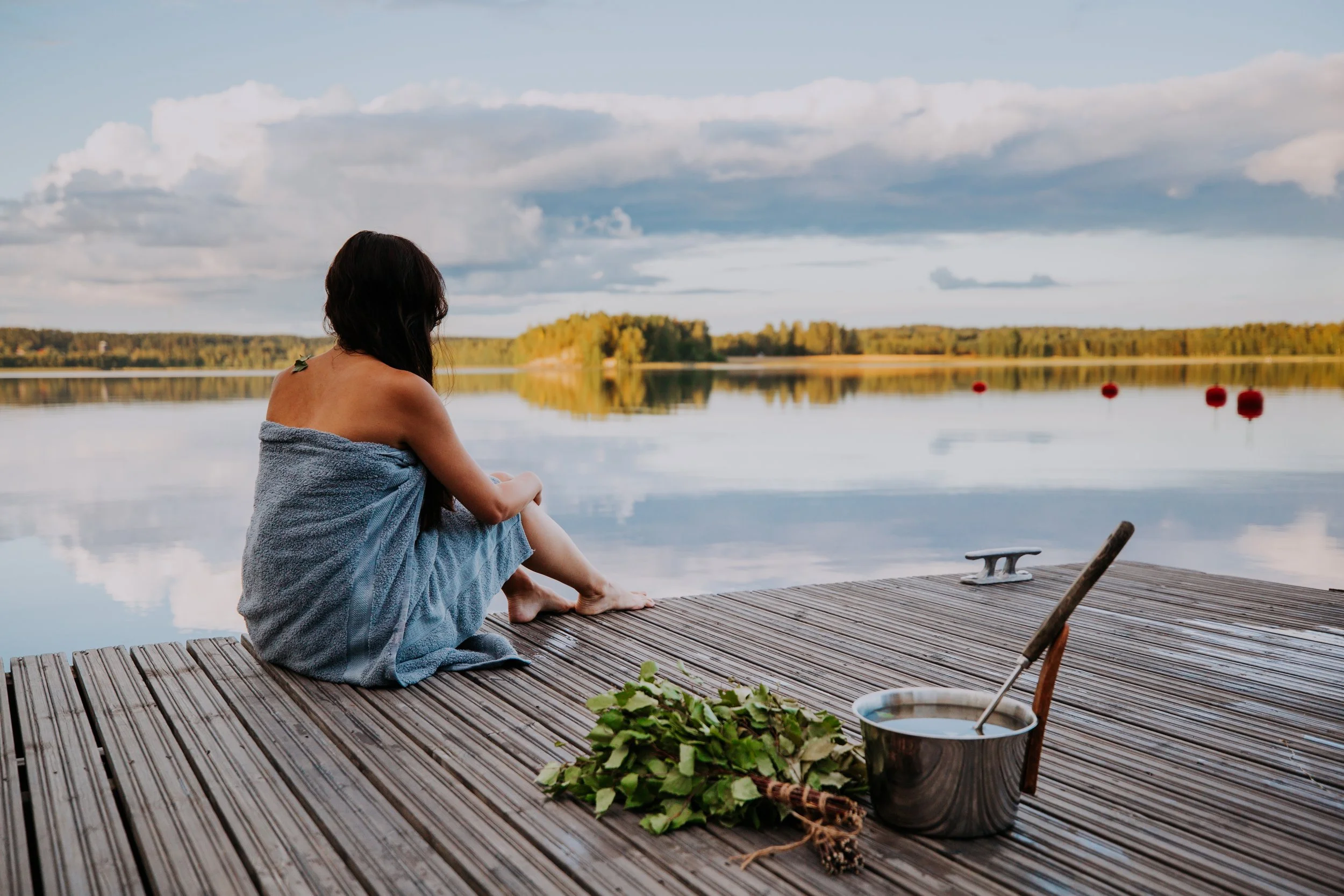 A person sitting on a wooden dock by a lake, wrapped in a towel, looking at the water and sky with cloudy weather. There are red buoys on the lake and green plants in a bowl on the dock.