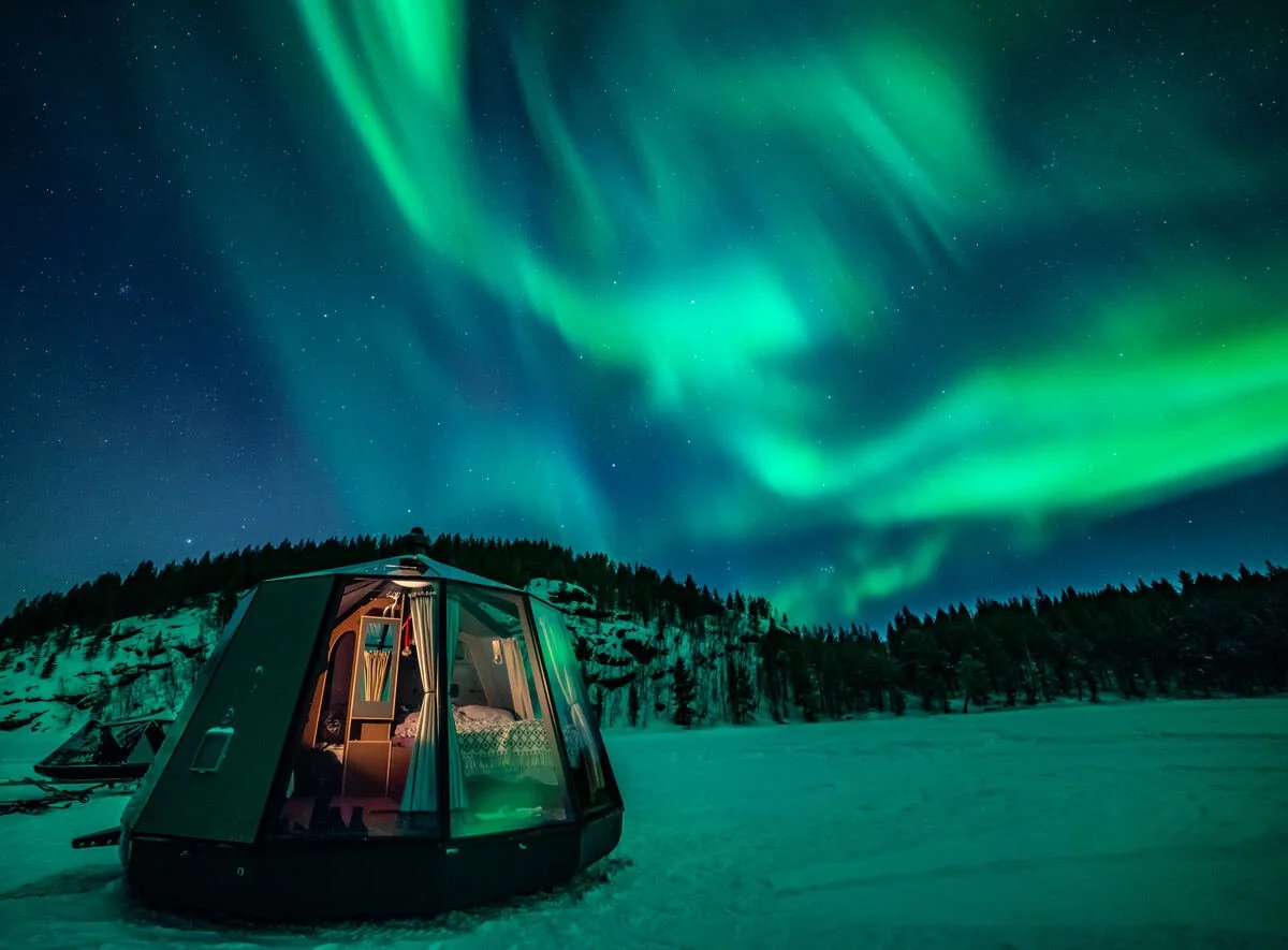 A glass-roofed Aurora Hut sits beneath vivid green Northern Lights in snowy Inari, Finnish Lapland.
