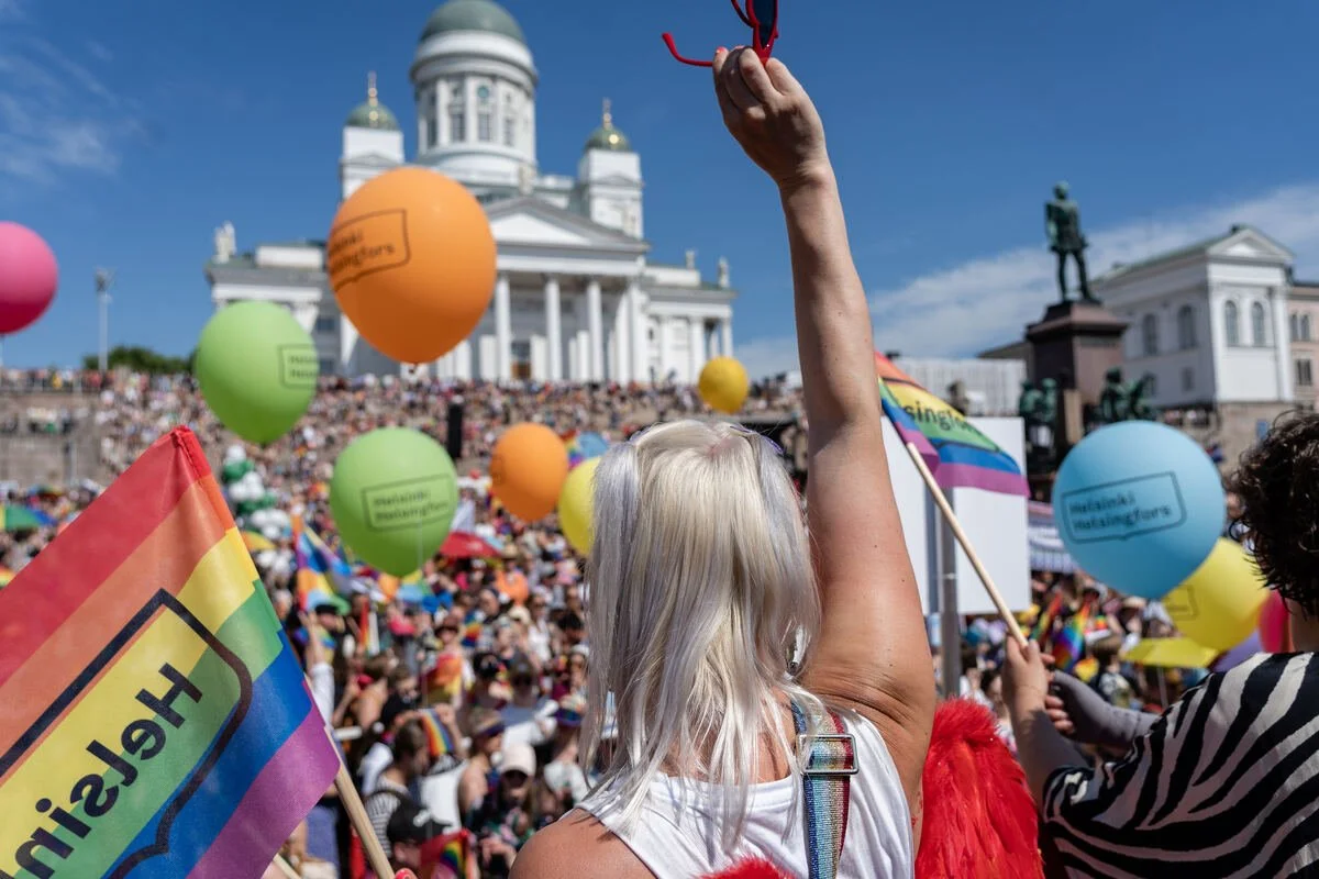 A large Helsinki Pride crowd with rainbow flags and colourful balloons gathers in front of Helsinki Cathedral.