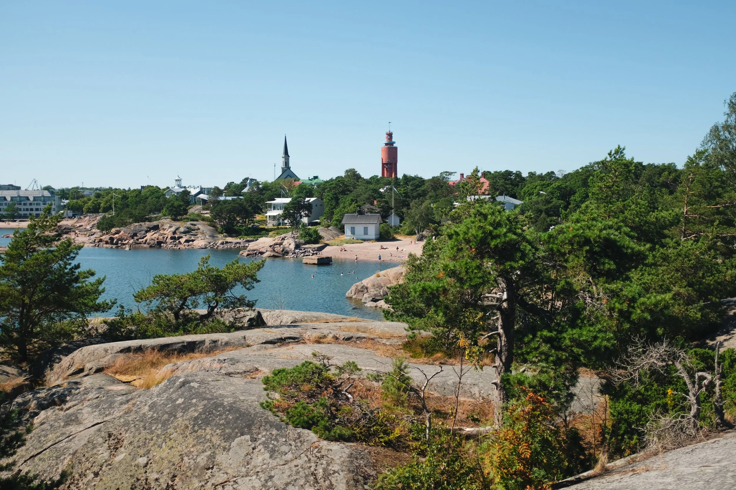 Scenic view of a waterfront of Hanko, Finland with trees, rocks, and houses, featuring a tall lighthouse and a church steeple against a clear blue sky.