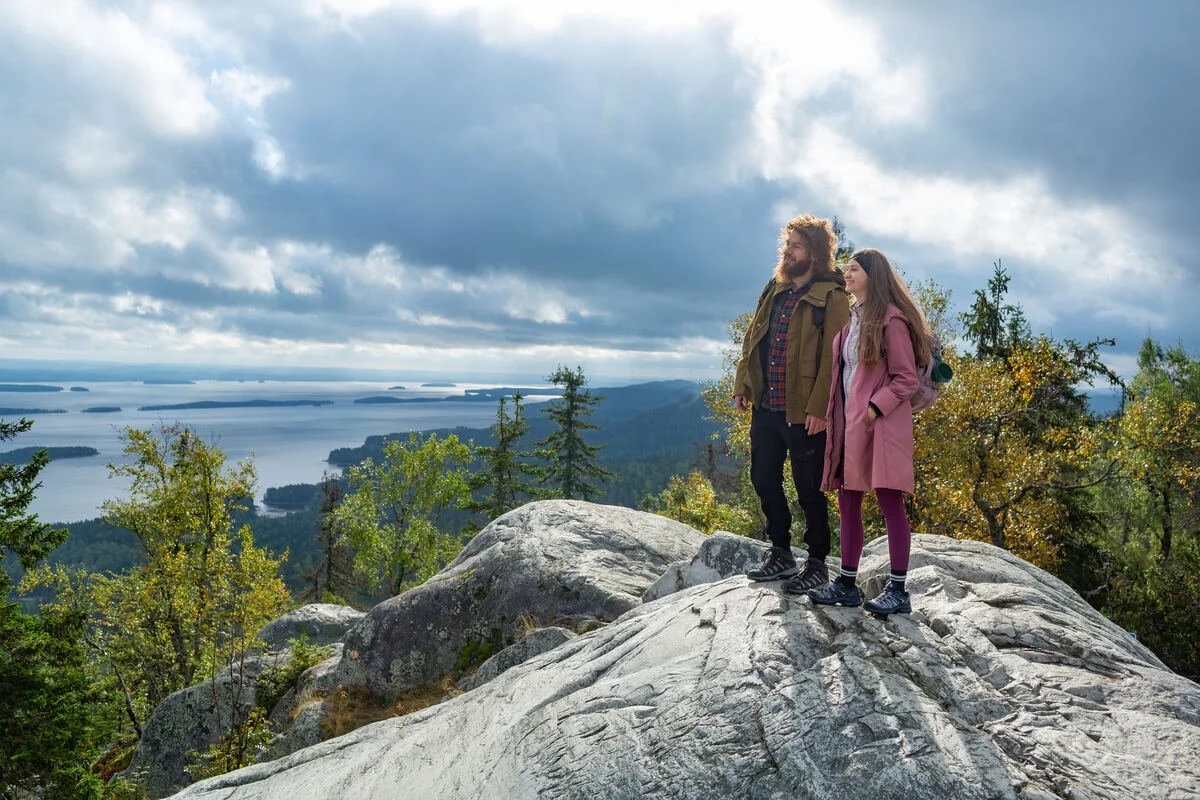 A couple stand on a rocky viewpoint overlooking lakes and forest at Koli National Park during autumn.