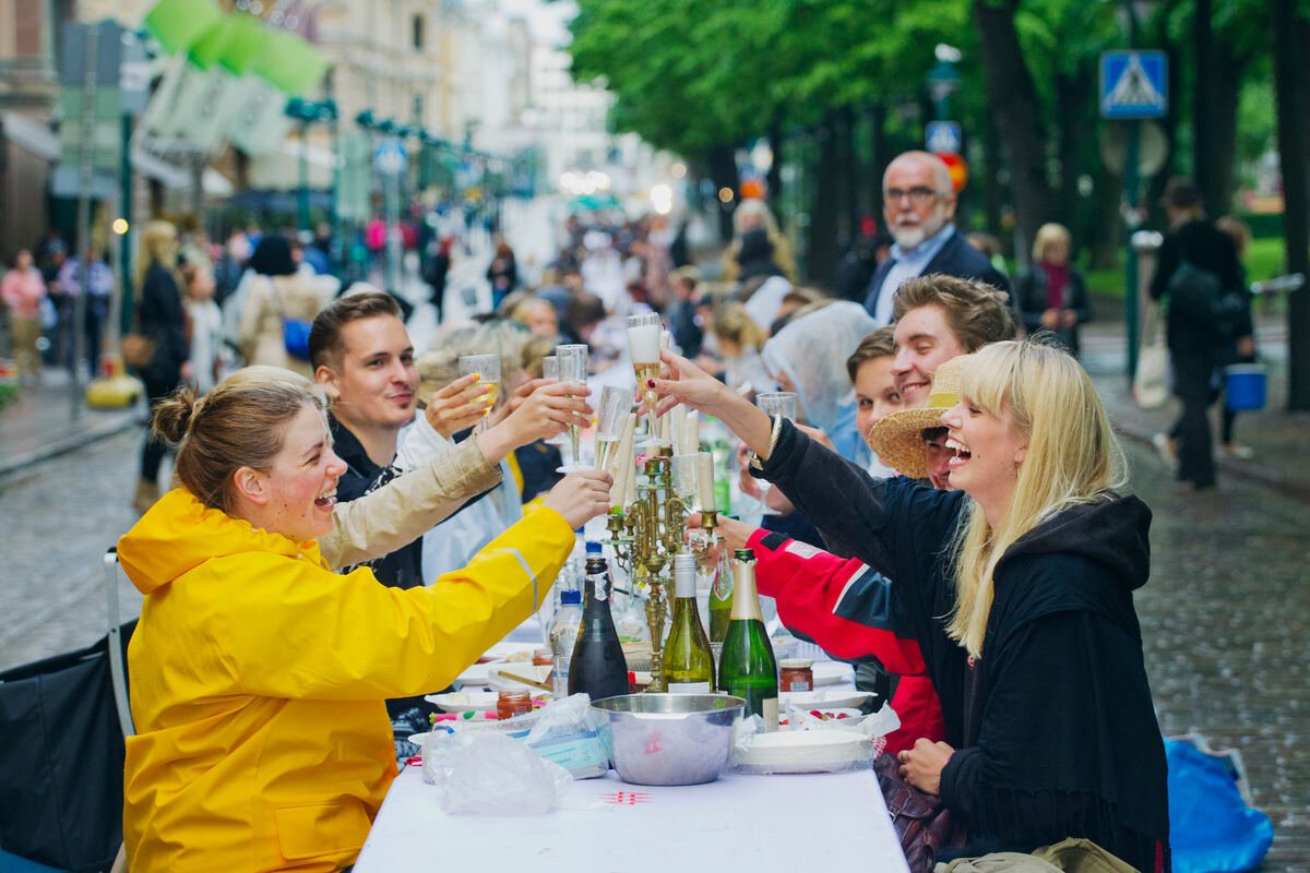 Friends raise glasses around a long outdoor dining table on a lively street in Helsinki.