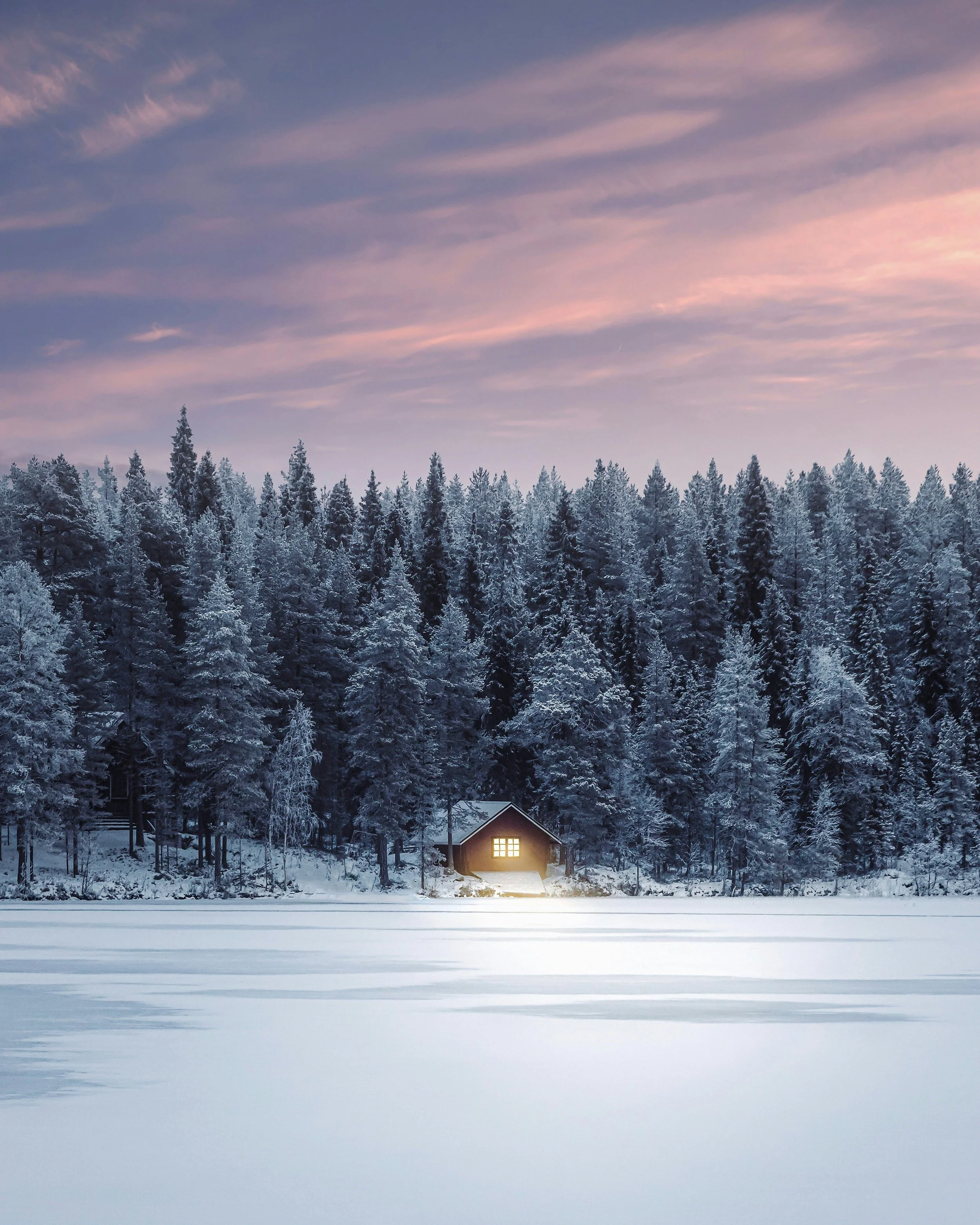 Cosy wooden cabin in Finland beside a frozen lake, surrounded by snow-covered pine forest beneath a pastel Nordic winter sky.