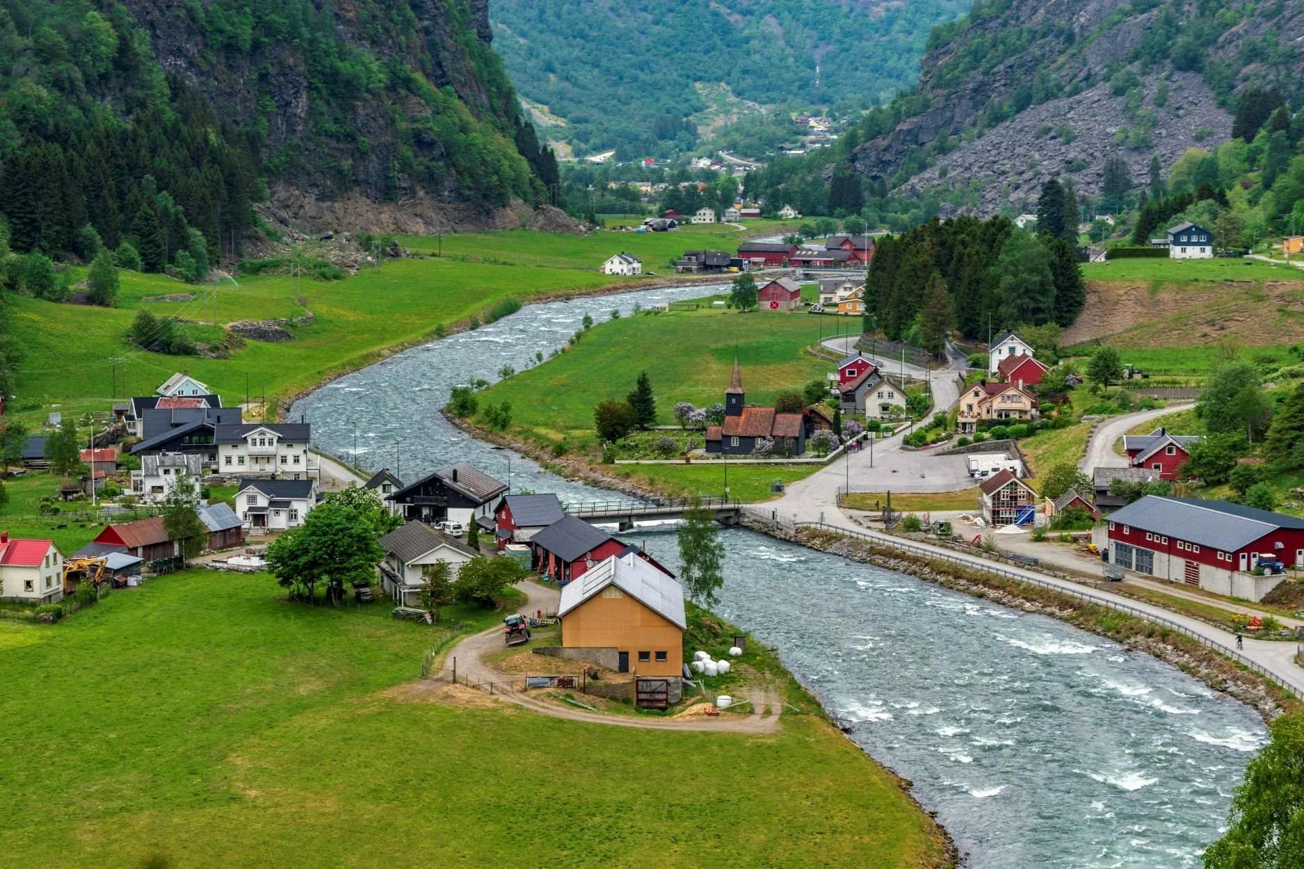 A scenic view of a small village nestled in a green, mountainous valley with a winding river flowing through it. The village has houses of various colors, including white, red, and yellow, surrounded by lush grassy fields and dense trees. The background features steep hills covered in forests and rocky terrain.