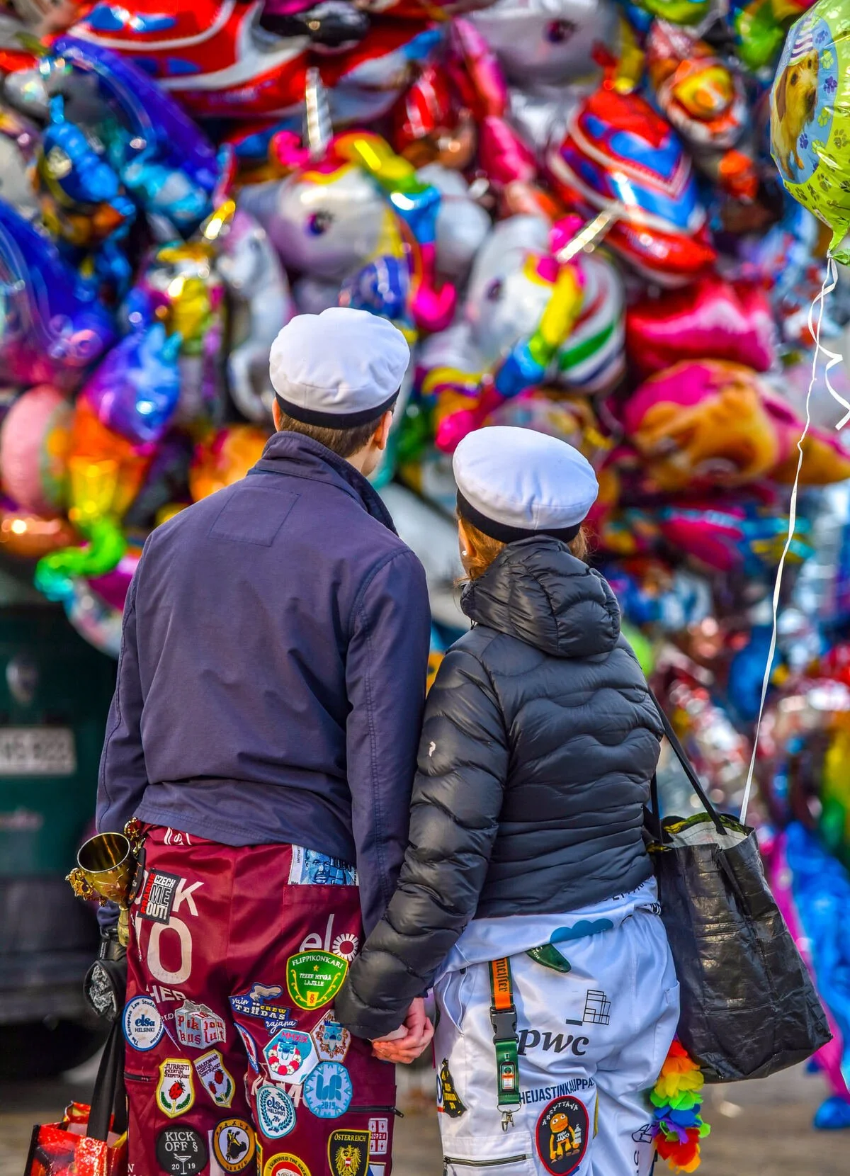 Two people wearing Finnish student caps stand in front of colourful balloons during Vappu celebrations in Helsinki.
