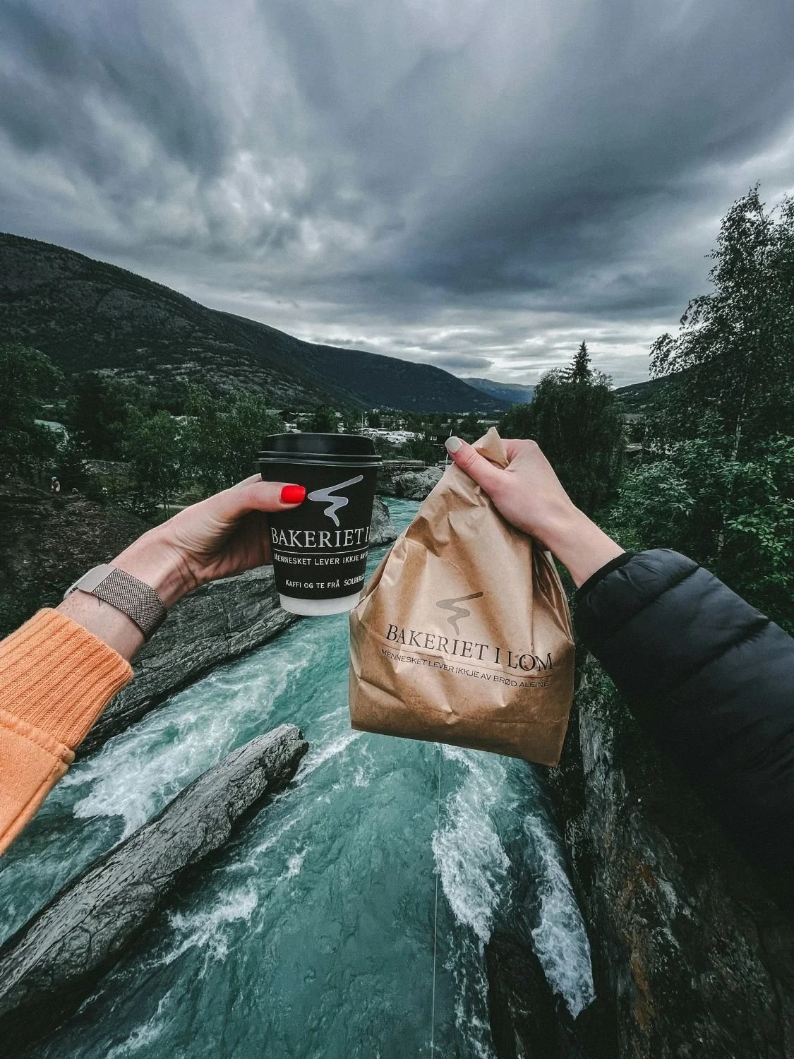 Coffee and a bakery bag from Bakeriet i Lom held above a rushing river surrounded by Norwegian mountains.