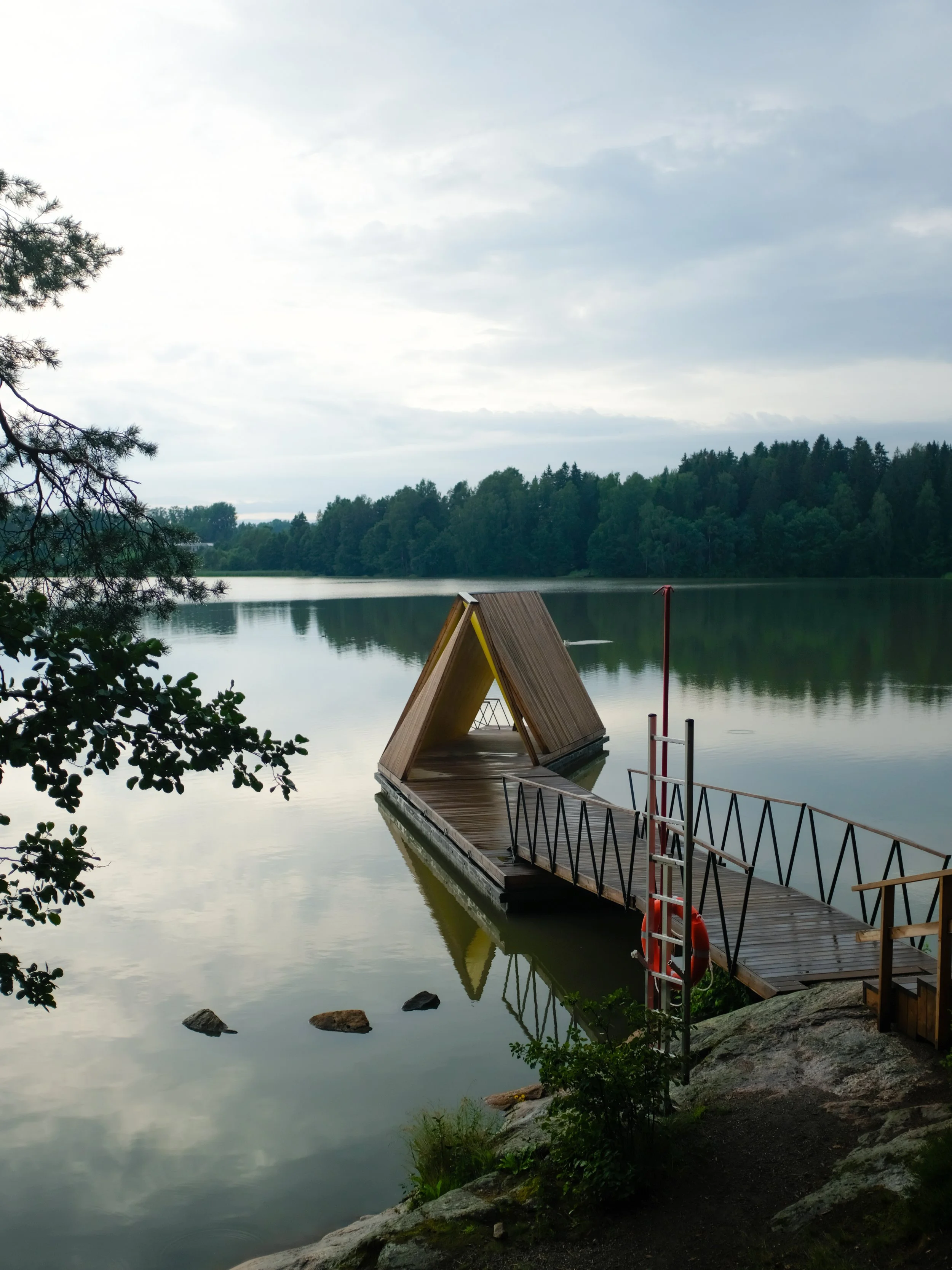 A wooden dock extends into a calm lake surrounded by green trees. There is an A-frame wooden structure on the dock, and the sky is overcast with clouds reflected on the water.