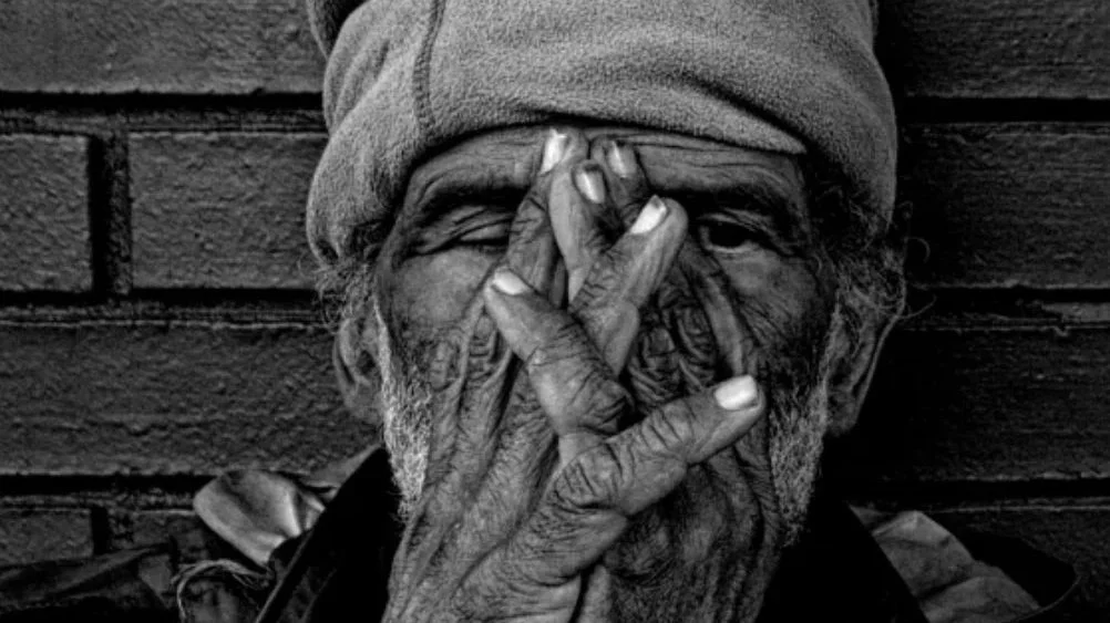 An elderly man with a beanie covering his head, covering part of his face with one hand, eyes closed, leaning against a brick wall.
