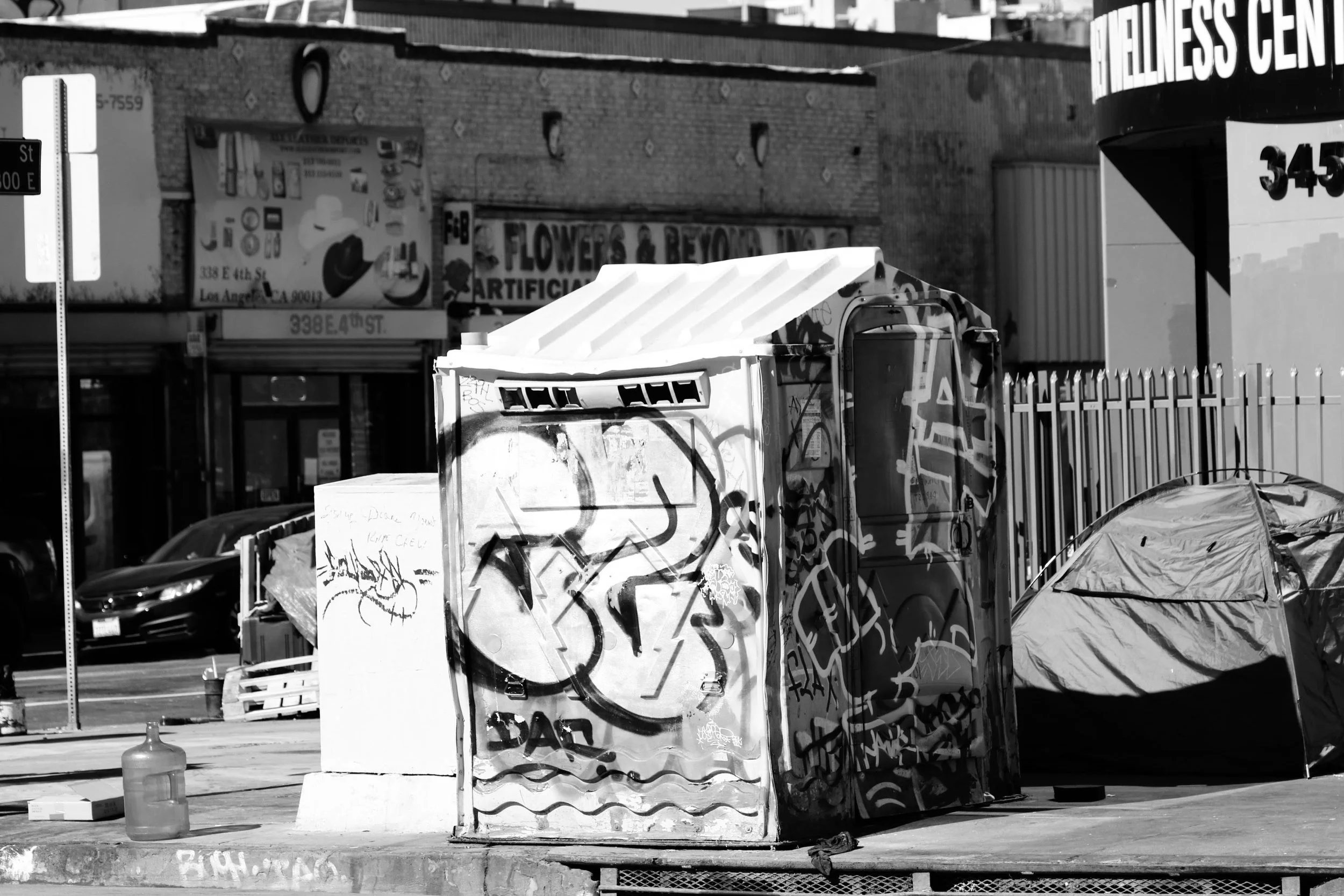 A graffiti-covered metal electrical box on a sidewalk, with a tent and a plastic bottle nearby, in an urban area with storefronts and parked cars in the background.