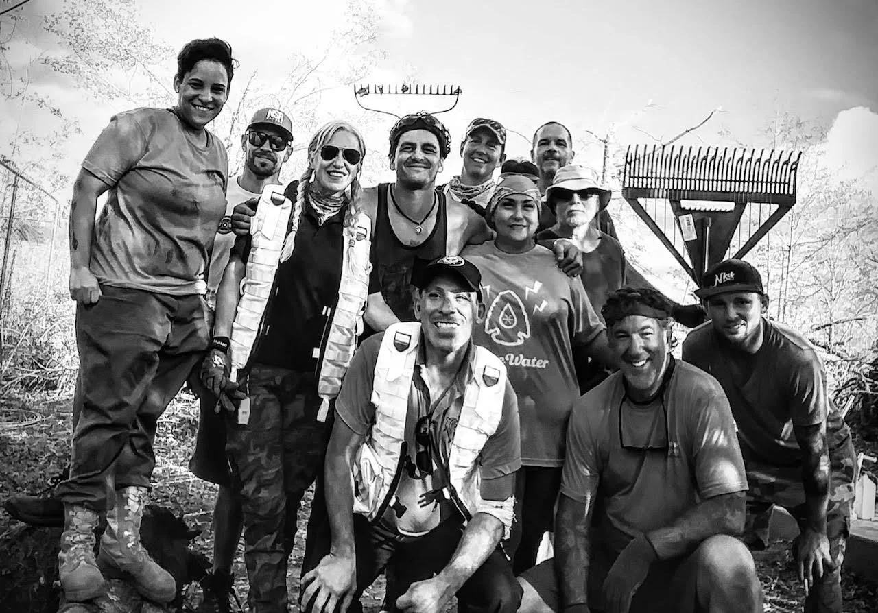 Group of people outdoors at a park, smiling for a photo, some wearing hats and sunglasses, with trees and a tall structure in the background.