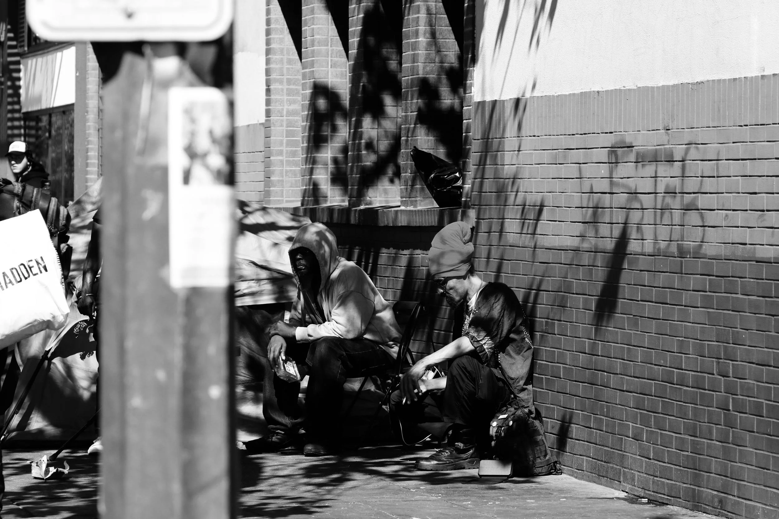Two men sit on chairs against a brick wall on a city sidewalk; one is wearing a hooded sweatshirt, and the other wears a turban and glasses, with bags placed beside them.