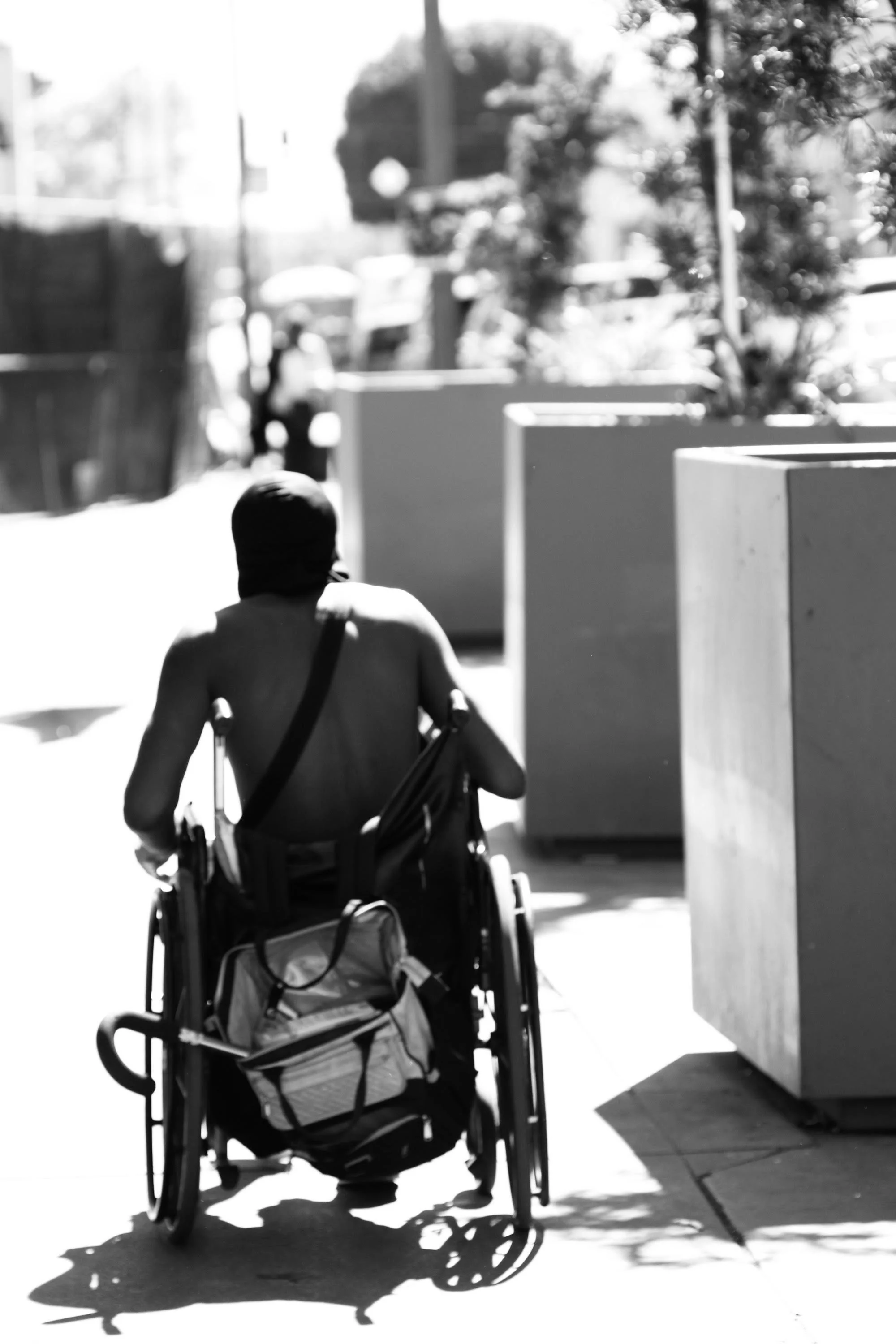 A person in a wheelchair with a backpack on the back, seen from behind, on a sidewalk with large planters and trees in an outdoor urban setting.