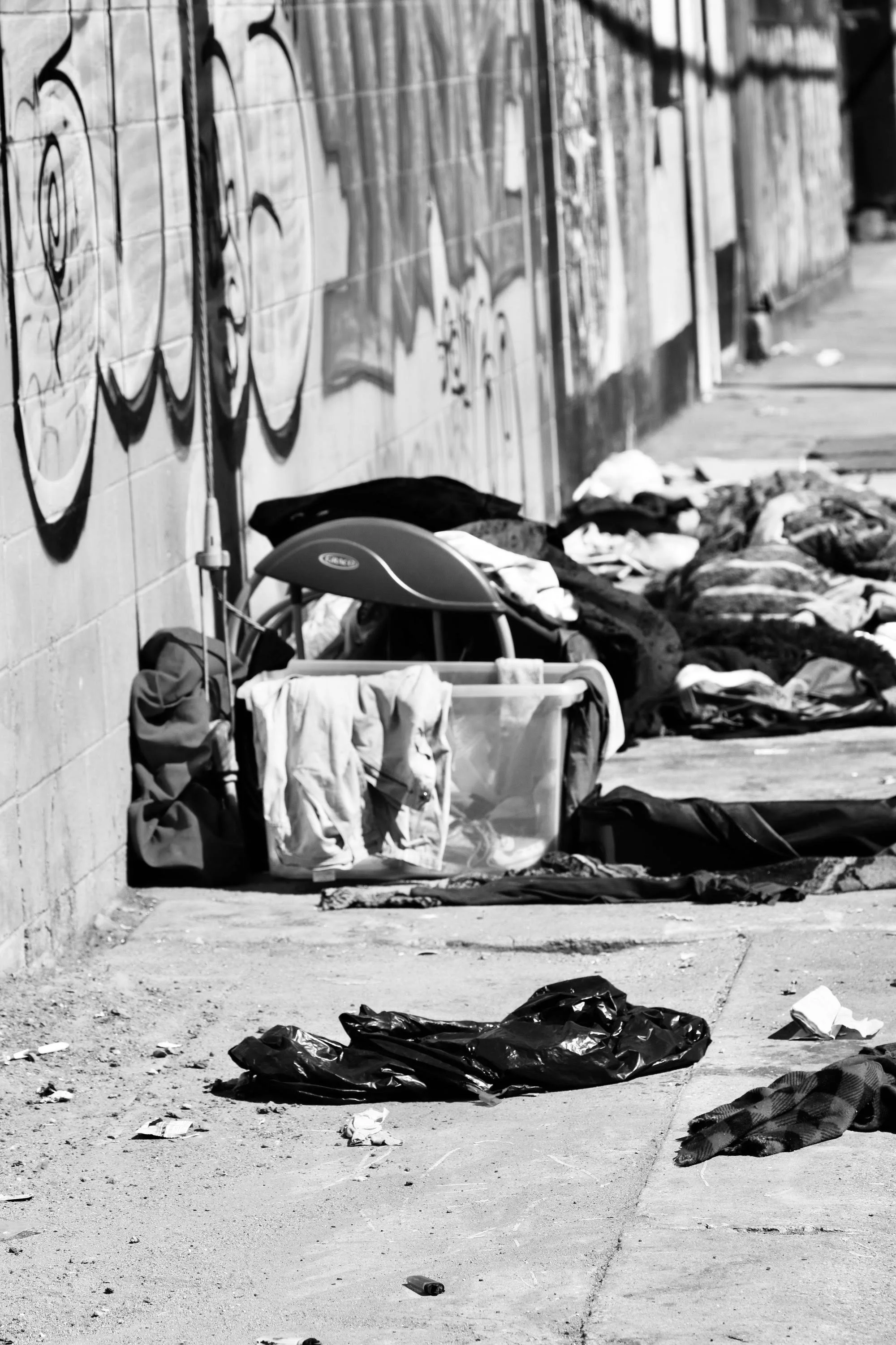 Litter and personal belongings, including clothing and bags, scattered on a sidewalk in front of a graffiti-covered wall.