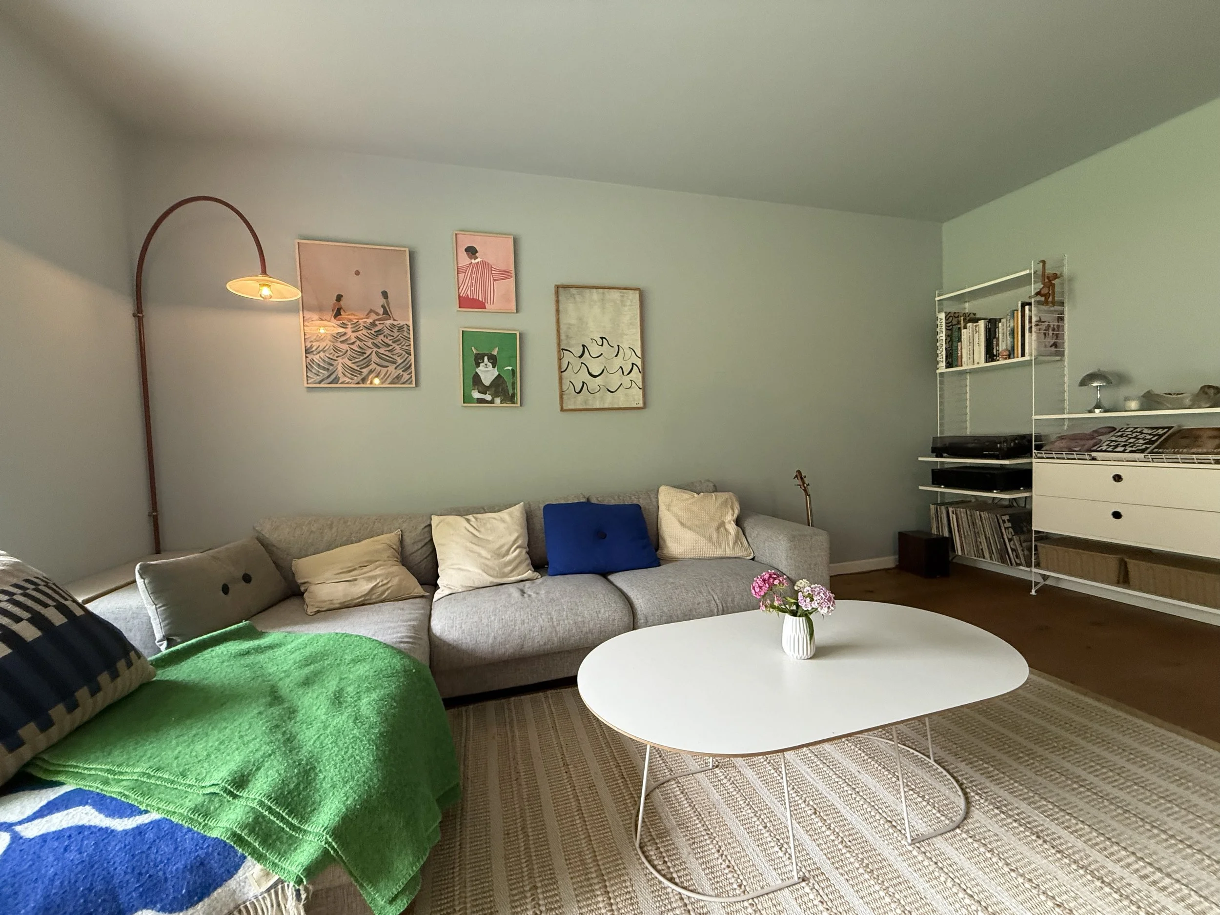 Wohnzimmer mit sanftem Blau-Grau, grauem Sofa, kräftigen grünen und blauen Akzenten und skandinavisch geprägter Einrichtung.