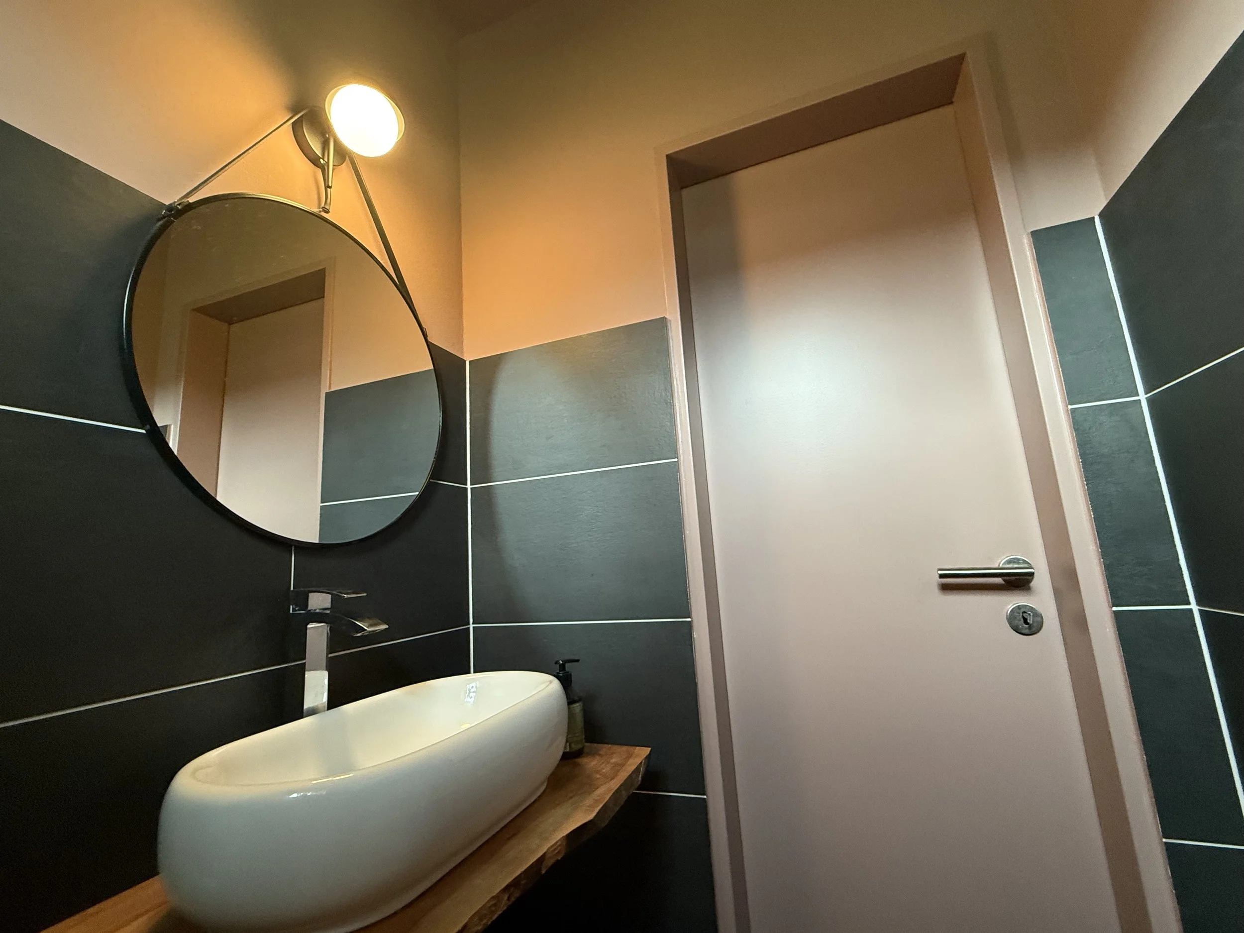Gäste-WC mit dunklen Wandfliesen, warmem Rosé und rundem Spiegel – kontrastreiches Farb- und Materialkonzept.