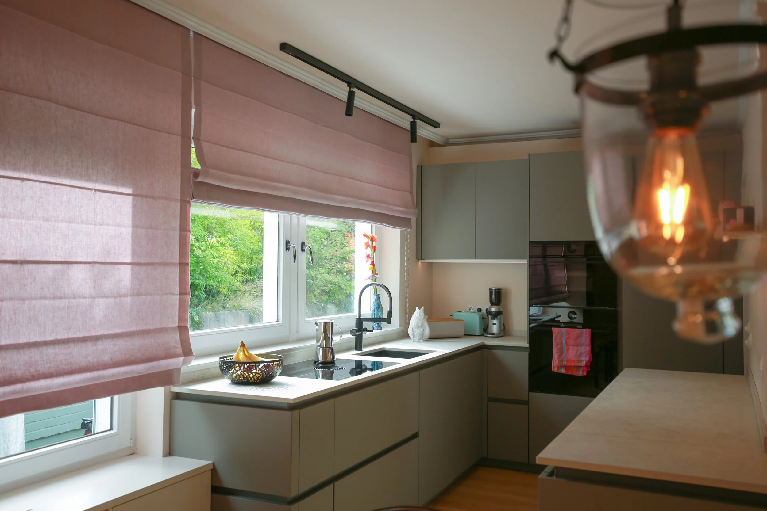 Individuelle Küchenplanung mit grauen Fronten, großzügiger Arbeitsfläche, rosa Raffrollos und warmem Holzboden.
