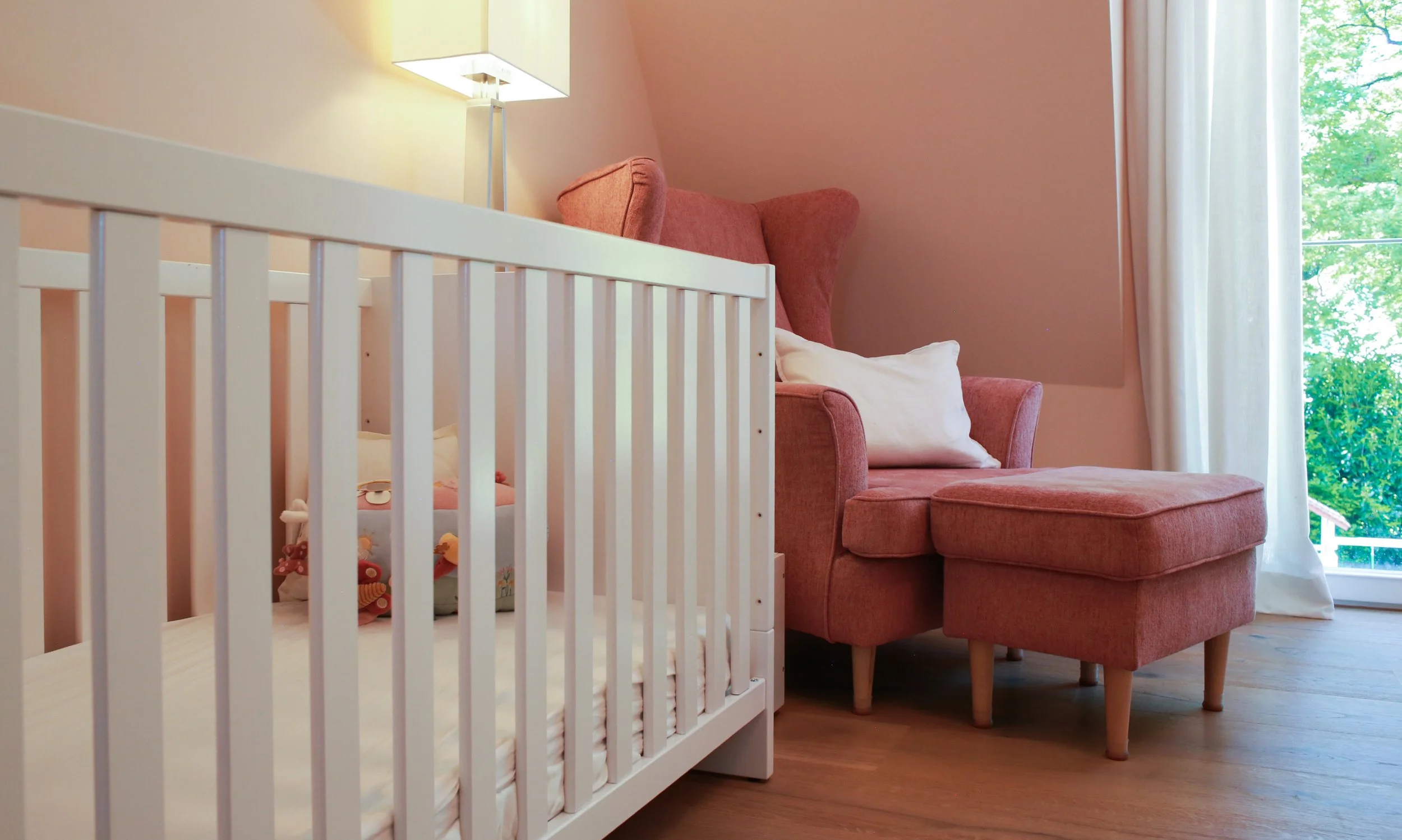 Kinderzimmer mit roséfarbenen Wänden und Dachschrägen, weißem Kinderbett und gemütlichem Sessel – warmes Farbkonzept für ein Familienhaus.
