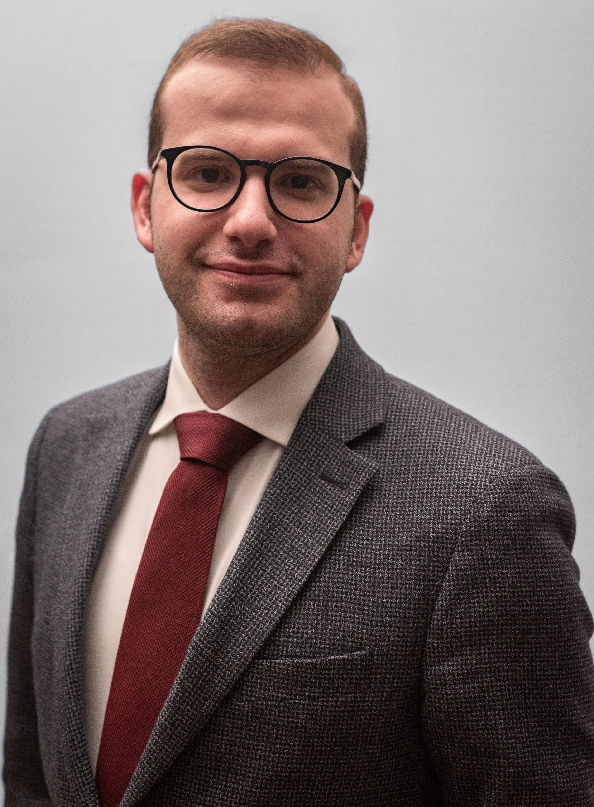 A young man with short brown hair, wearing black-rimmed glasses, a white shirt, a red tie, and a gray checkered blazer, standing against a light gray background.