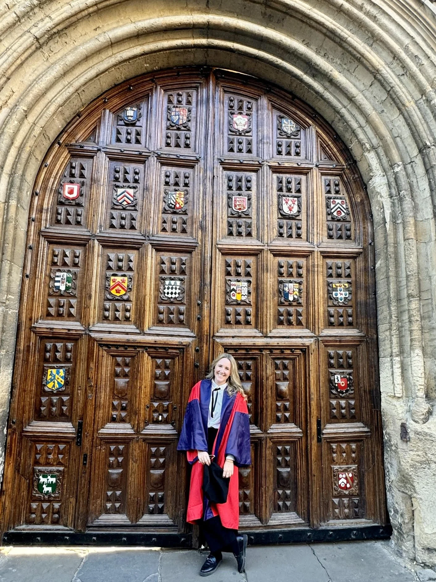 A woman in academic regalia standing in front of a large wooden door with decorative square panels, each featuring a colorful heraldic shield or coat of arms. The door is arched and set into stone architecture.