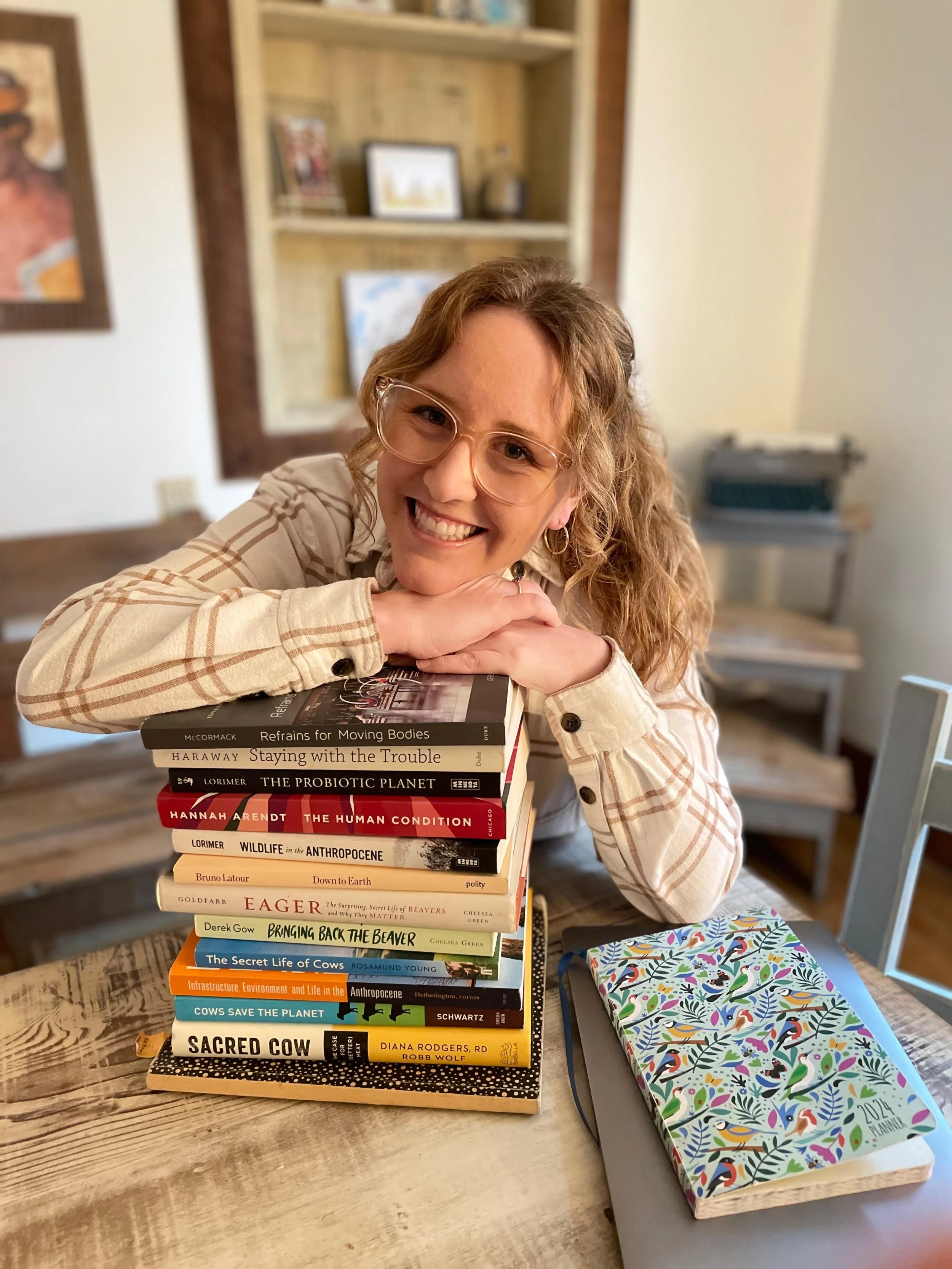 A woman with glasses and curly hair smiling and resting her chin on a stack of books on a wooden table. Behind her, a bookshelf and some framed pictures are visible. There is also a colorful notebook on the table.