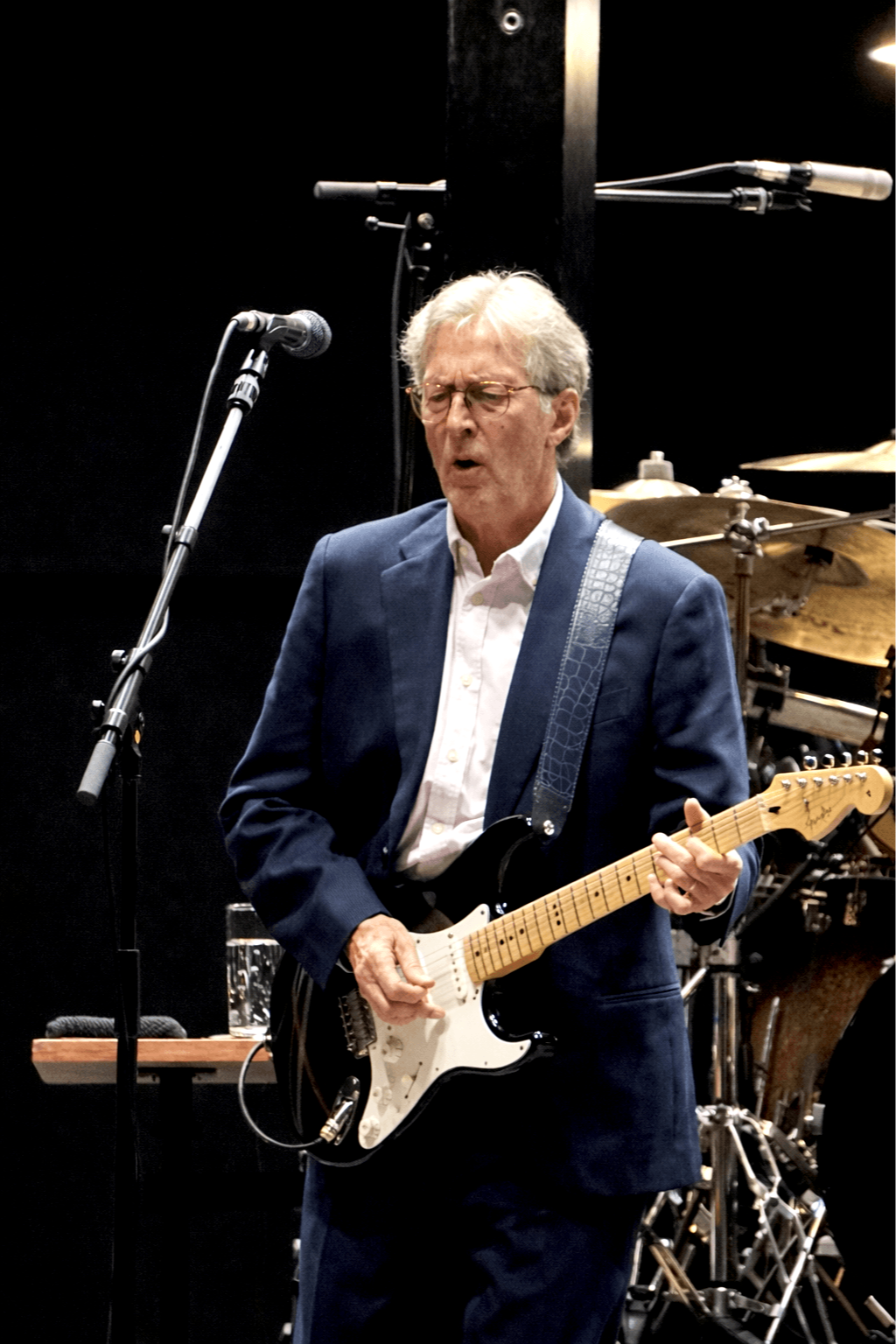 Eric Clapton in a navy blue suit, and a white shirt, playing an electric guitar on The Royal Albert Hall stage in May 2025. There is a microphone in front of him, and drums are visible in the background.