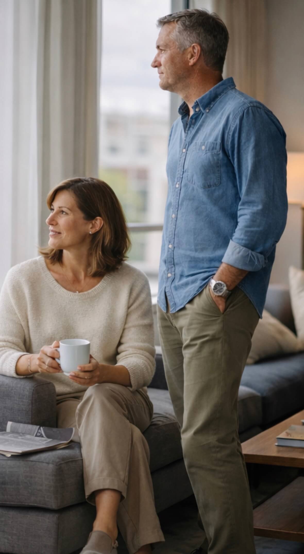 A middle-aged woman with shoulder-length brown hair sitting on a gray sofa holding a white mug, looking up and smiling while a middle-aged man with gray hair and beard, wearing a blue denim shirt and beige pants, stands beside her with his hands in his pockets, looking out the window of a modern, well-lit living room.